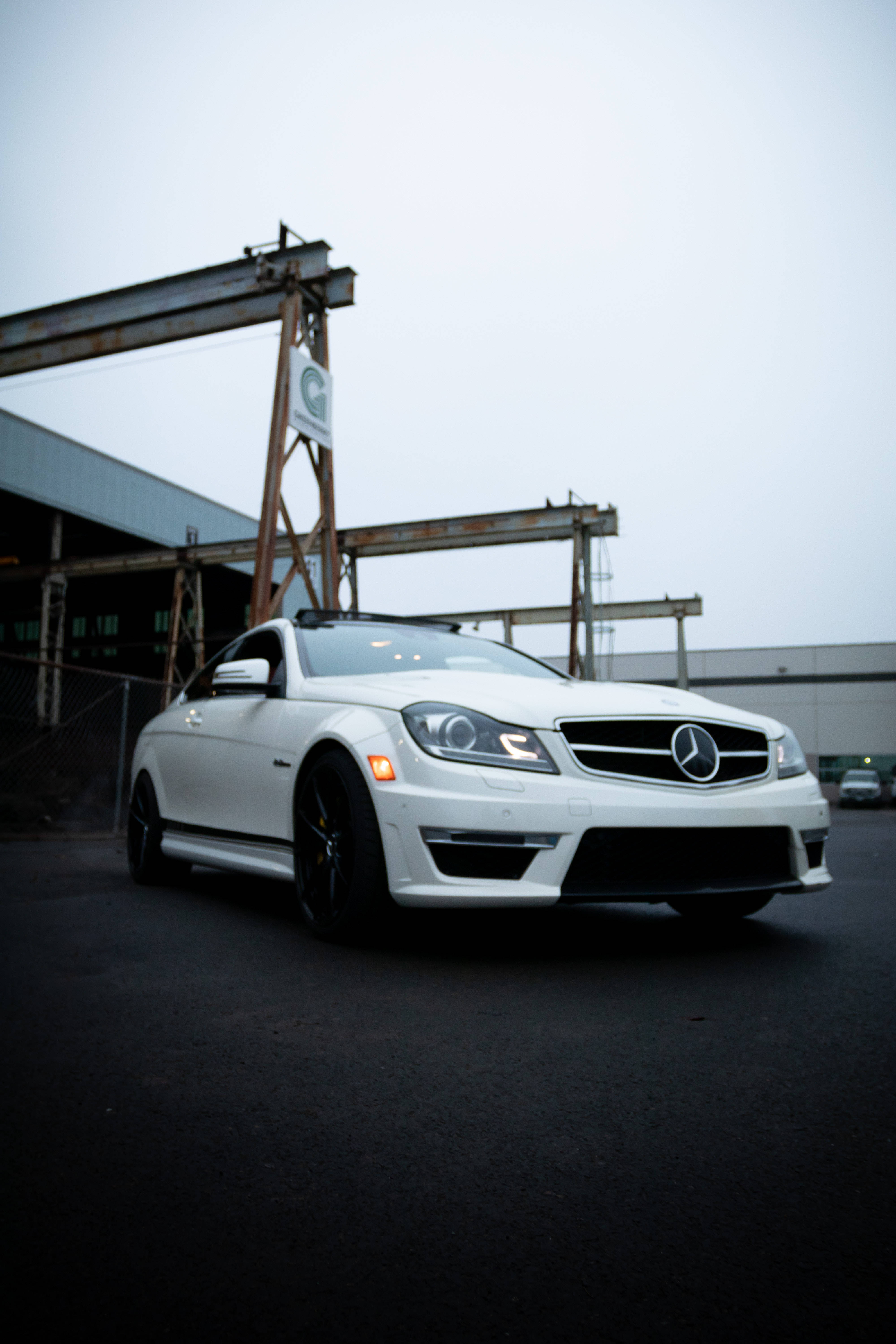 白色梅賽德斯-AMG iPhone 壁紙 — 一架時尚的白色平治跑車停泊在工業起重機前，天空多雲。