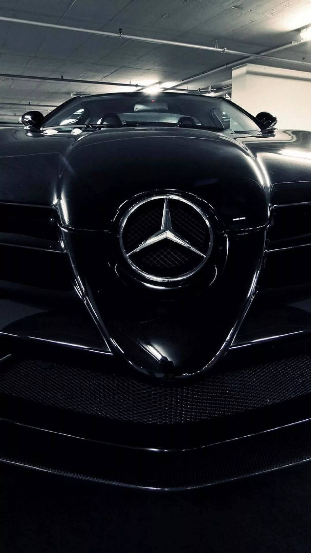 梅賽德斯-AMG車標保險桿 iPhone壁紙 壁紙 — 一輛時尚的黑色梅賽德斯跑車在刺眼的車庫燈光下閃閃發光，配備碳纖維格柵和標誌性的星形徽章。