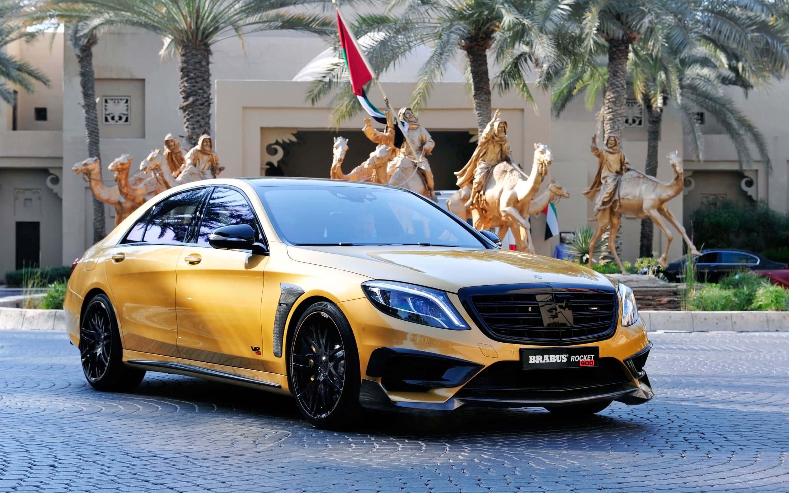 金色梅賽德斯-奔馳AMG S65 壁紙 — 一輛閃亮的金色Brabus Rocket 900轎車停在鵝卵石上，前方有金色駱駝雕像和棕櫚樹。