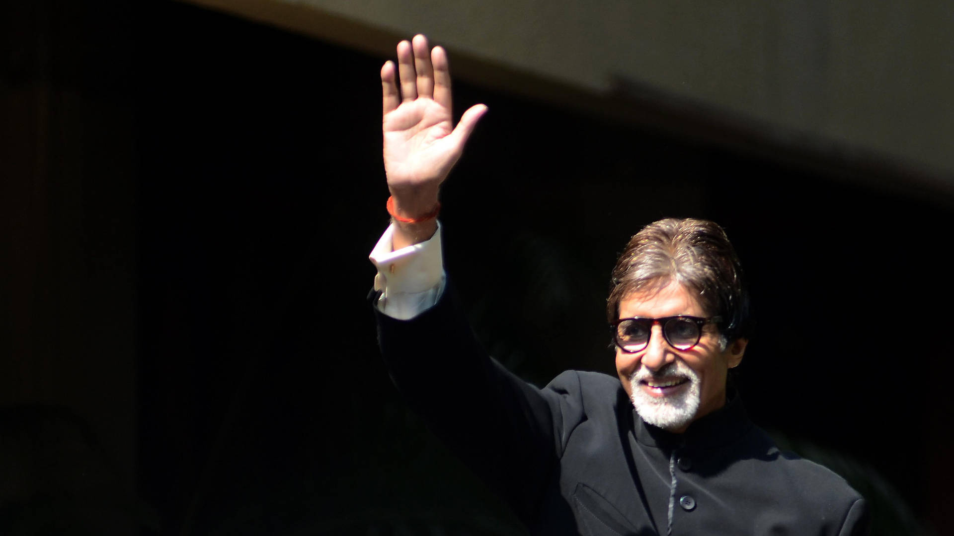 電影製片人 Amitabh Bachchan 壁紙 — 一位戴眼鏡、留白鬍子的微笑男士在明亮陽光下，對著深色背景愉快地揮手。