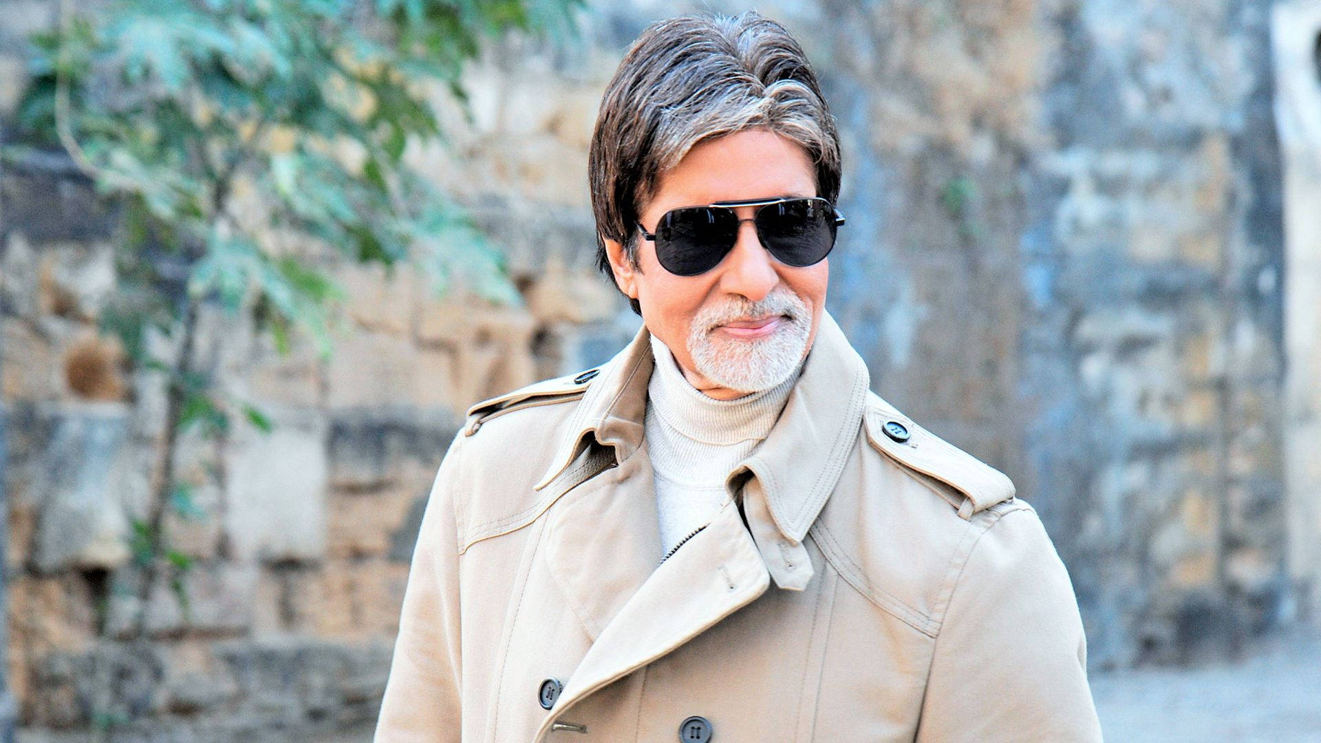 現代風 Amitabh Bachchan 壁紙 — 一位留著灰色鬍鬚、戴著太陽眼鏡的時尚男士，身穿米色風衣，背景是石牆。