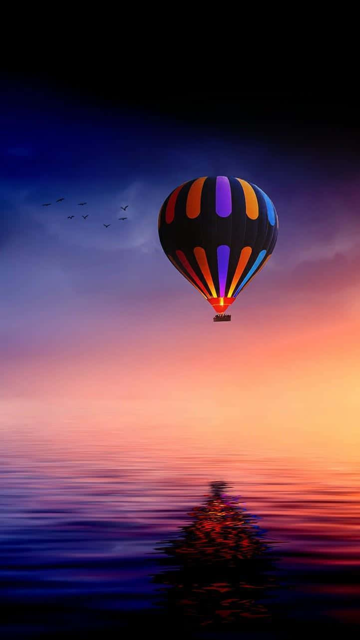 AMOLED S熱氣球日落海景 壁紙 — 一個色彩繽紛的熱氣球在日落時分漂浮在平靜的水面上，其倒影在下方閃爍，鳥兒在頭頂飛翔。