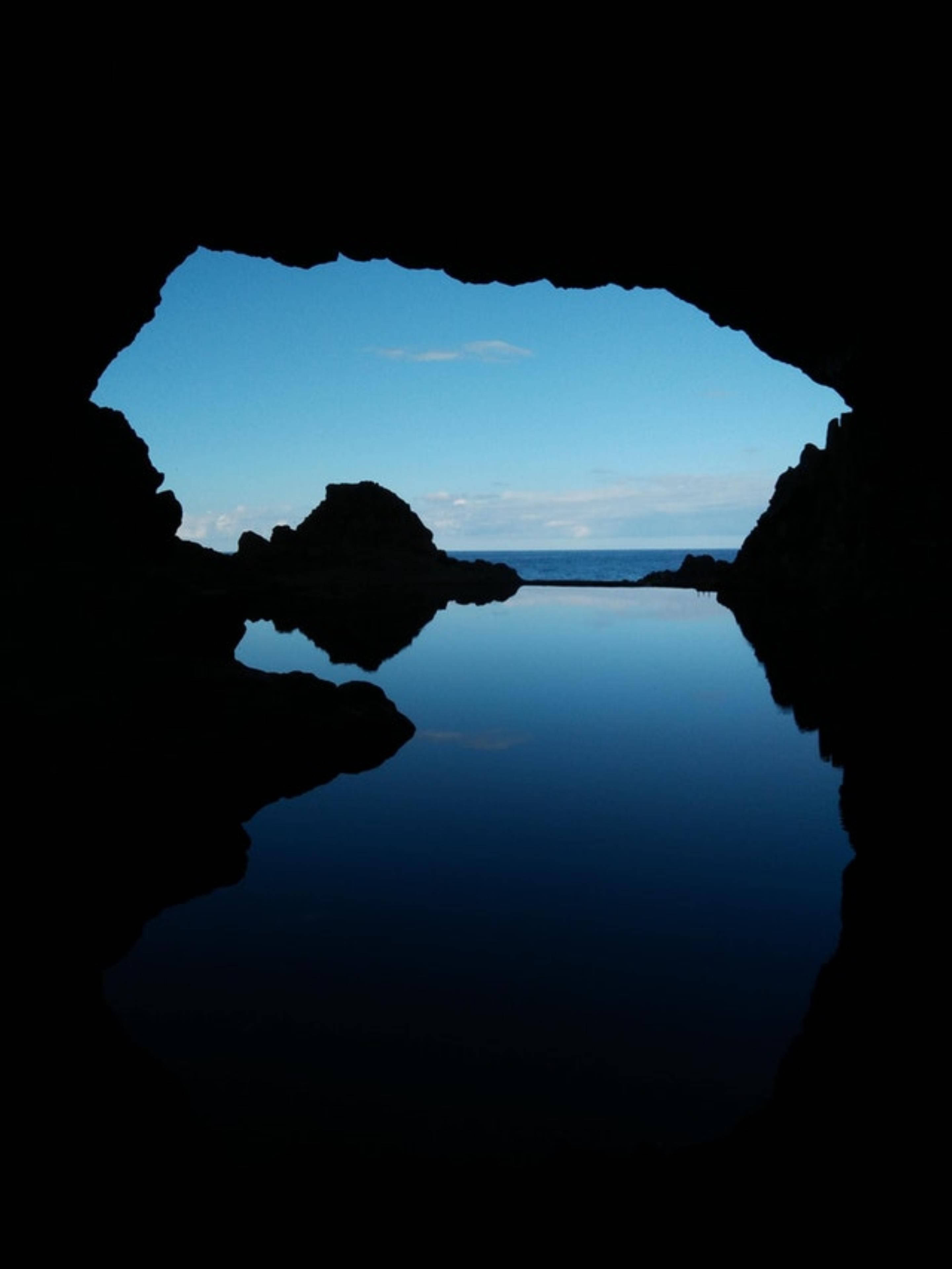 AMOLED 洞穴海水 壁紙 — 一個黑暗的洞穴框住了一片寧靜的藍天和寧靜的海洋，完美地倒映在下面的靜水中。
