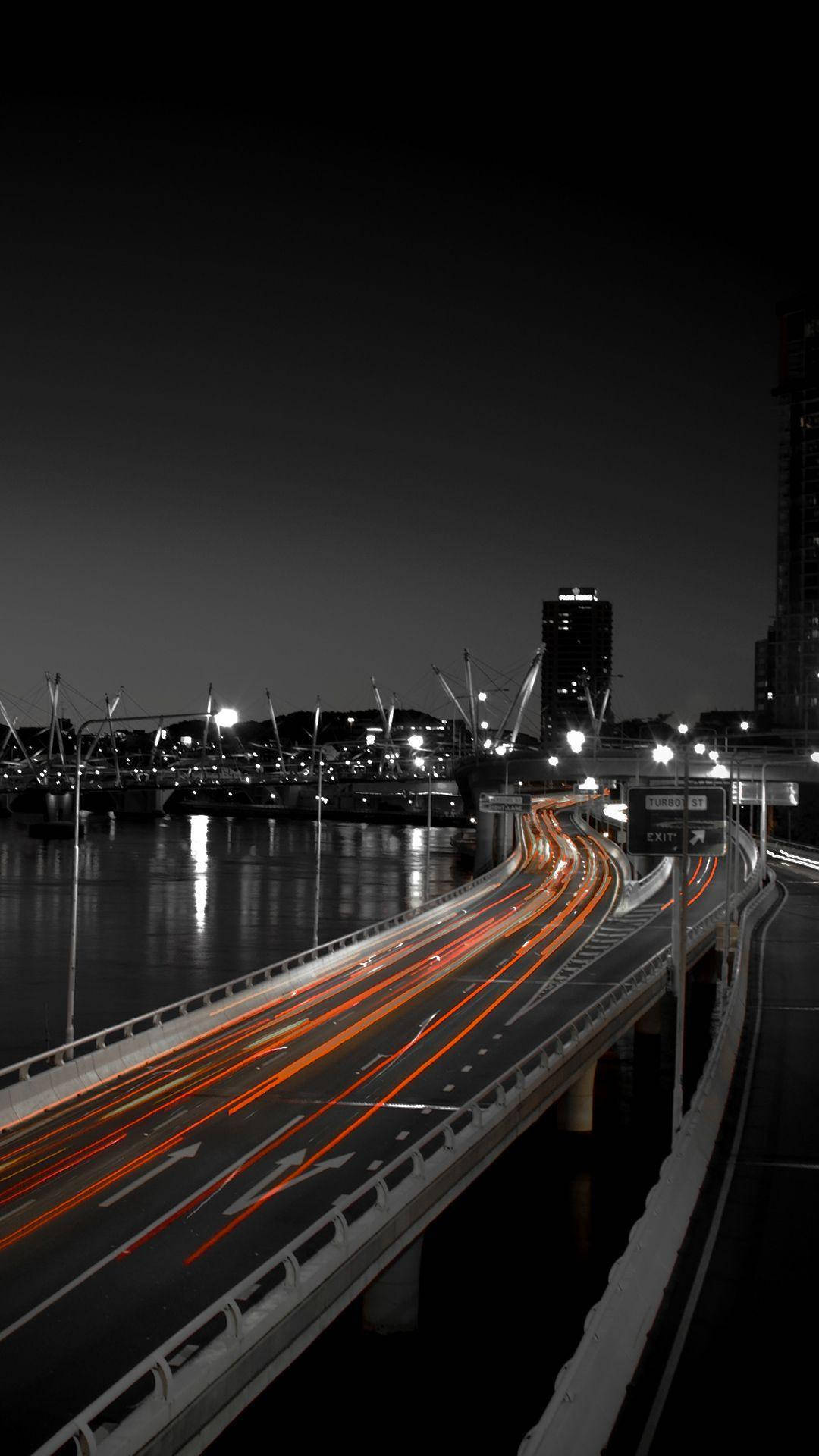 AMOLED 長曝光城市夜景 壁紙 — 長時間曝光捕捉到夜間橋樑上流動的橙色發光交通軌跡，背景是黑暗的水面和城市燈光。