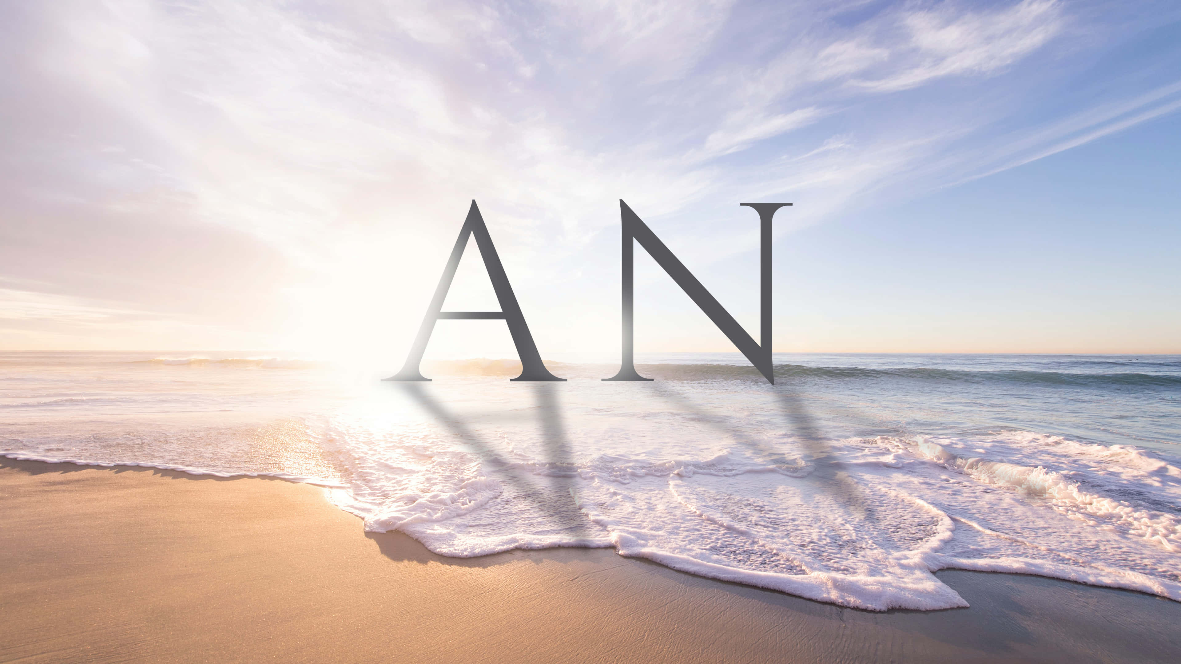 「AN」立於海洋邊 壁紙 — 大型金屬字母AN在陽光明媚的海灘上，日出時海洋波浪輕輕拍打其底部。