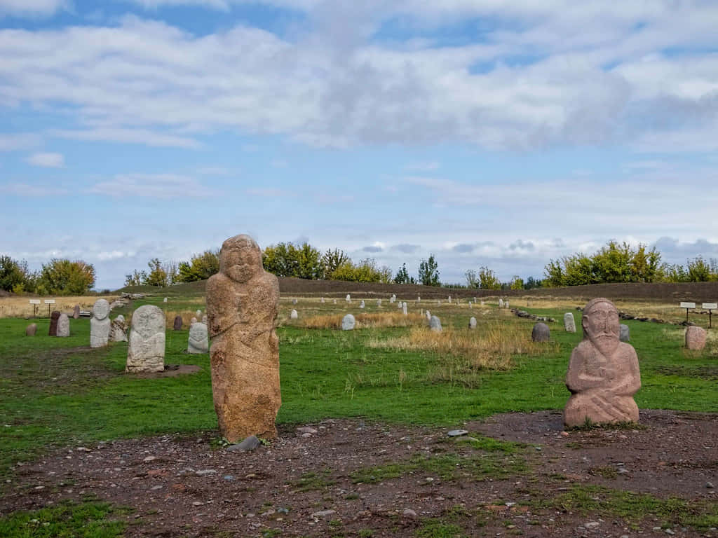 Kyrgyzstan 桌布 壁紙 — 古代石像莊嚴地矗立在草地上，天空部分多雲，遠處有秋天的樹木。