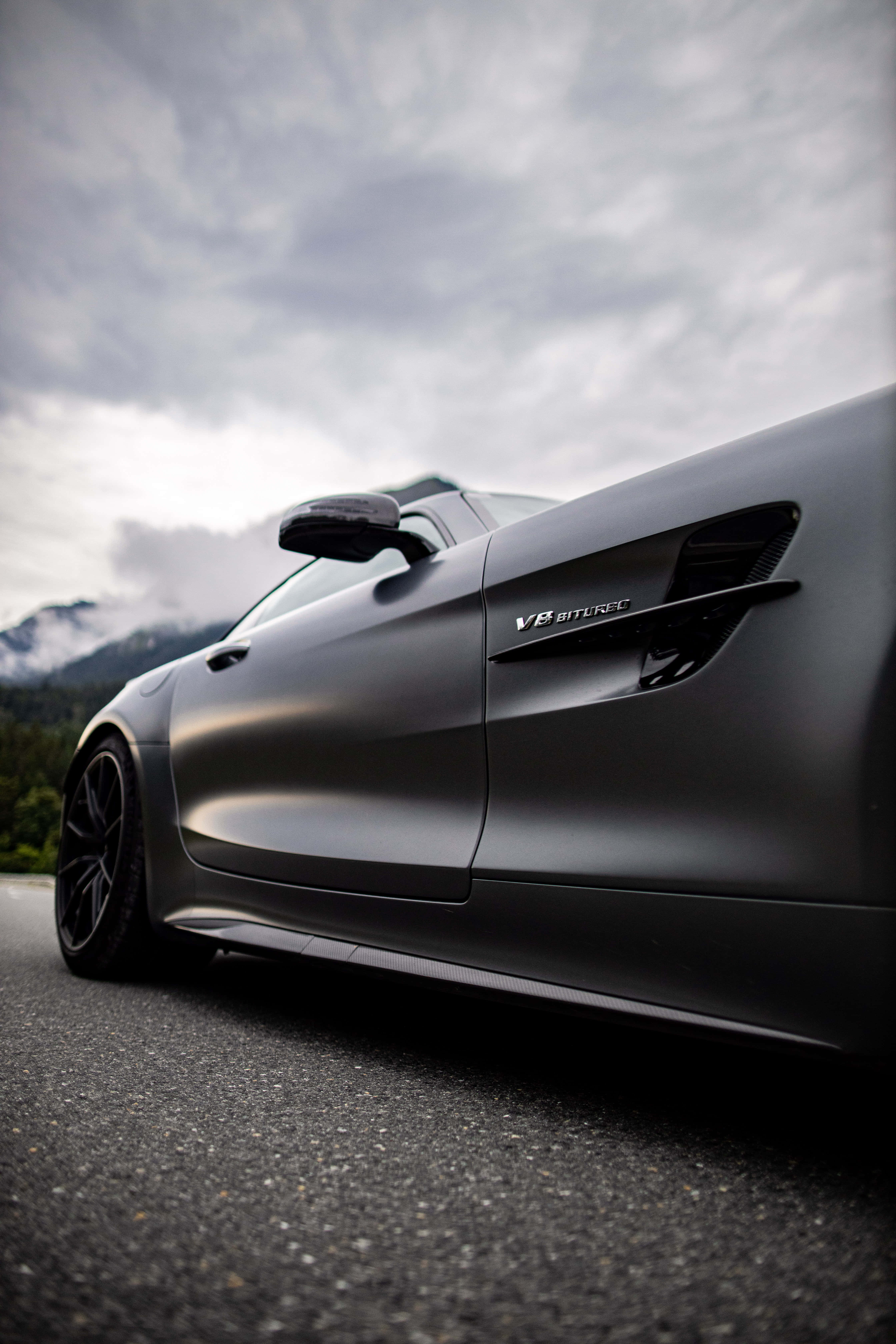 美學Android手機背景：AMG GT-R 車款 壁紙 — 一輛帶有V8 BITURBO徽章的啞光黑色跑車停在瀝青路上，背景是陰天和霧氣繚繞的山脈。