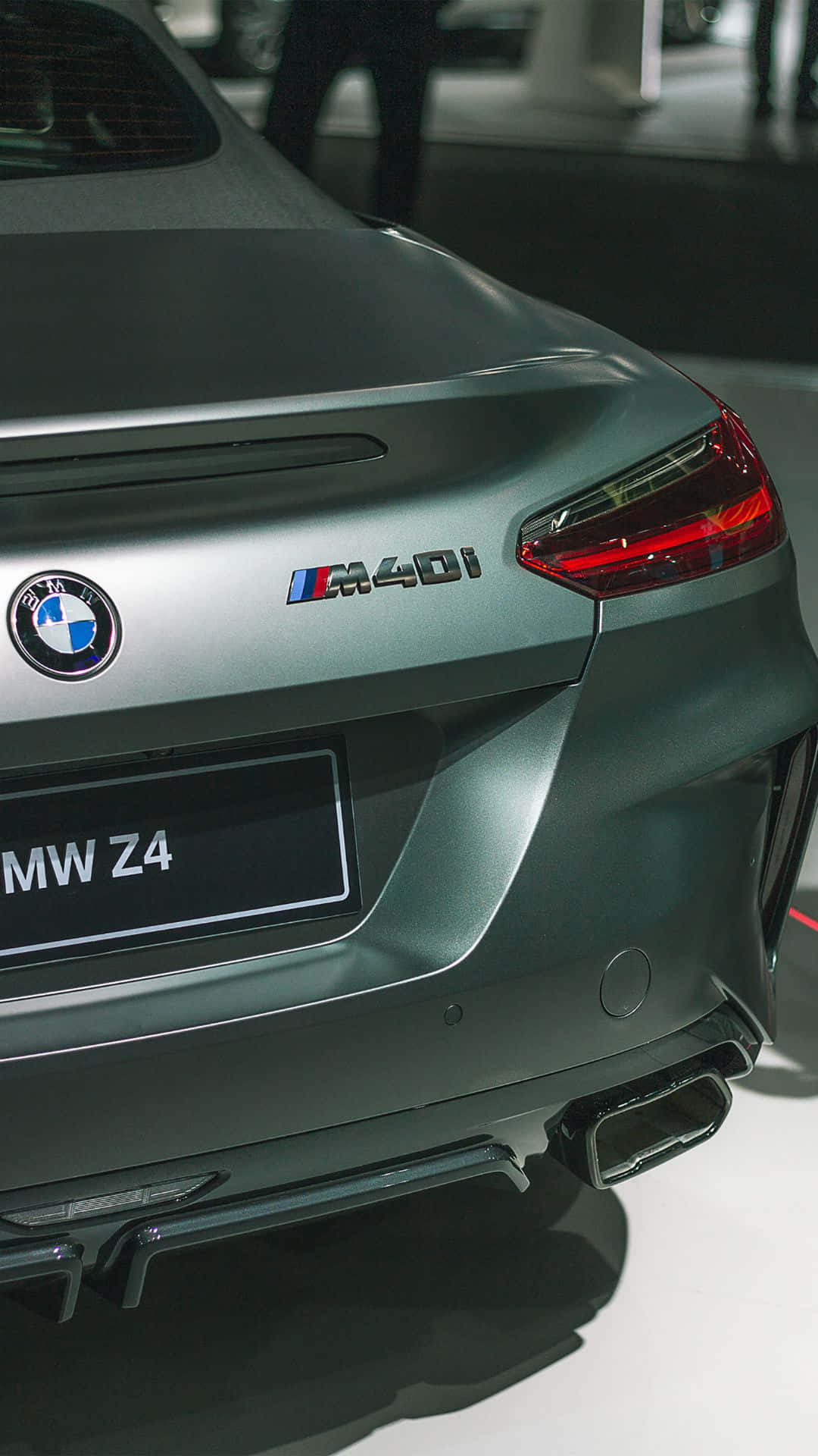 Android BMW 全配最新科技 壁紙 — 啞光灰色BMW Z4 M40i跑車尾部特寫，在明亮的展廳燈光下展示時尚尾燈和雙排氣管。