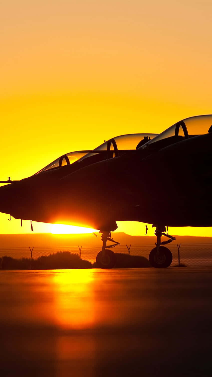 黃昏機場停機坪Android巨型客機背景 壁紙 — 戰鬥機的剪影矗立在跑道上，夕陽在它們身後以火熱的橙色和黃色色調落下。