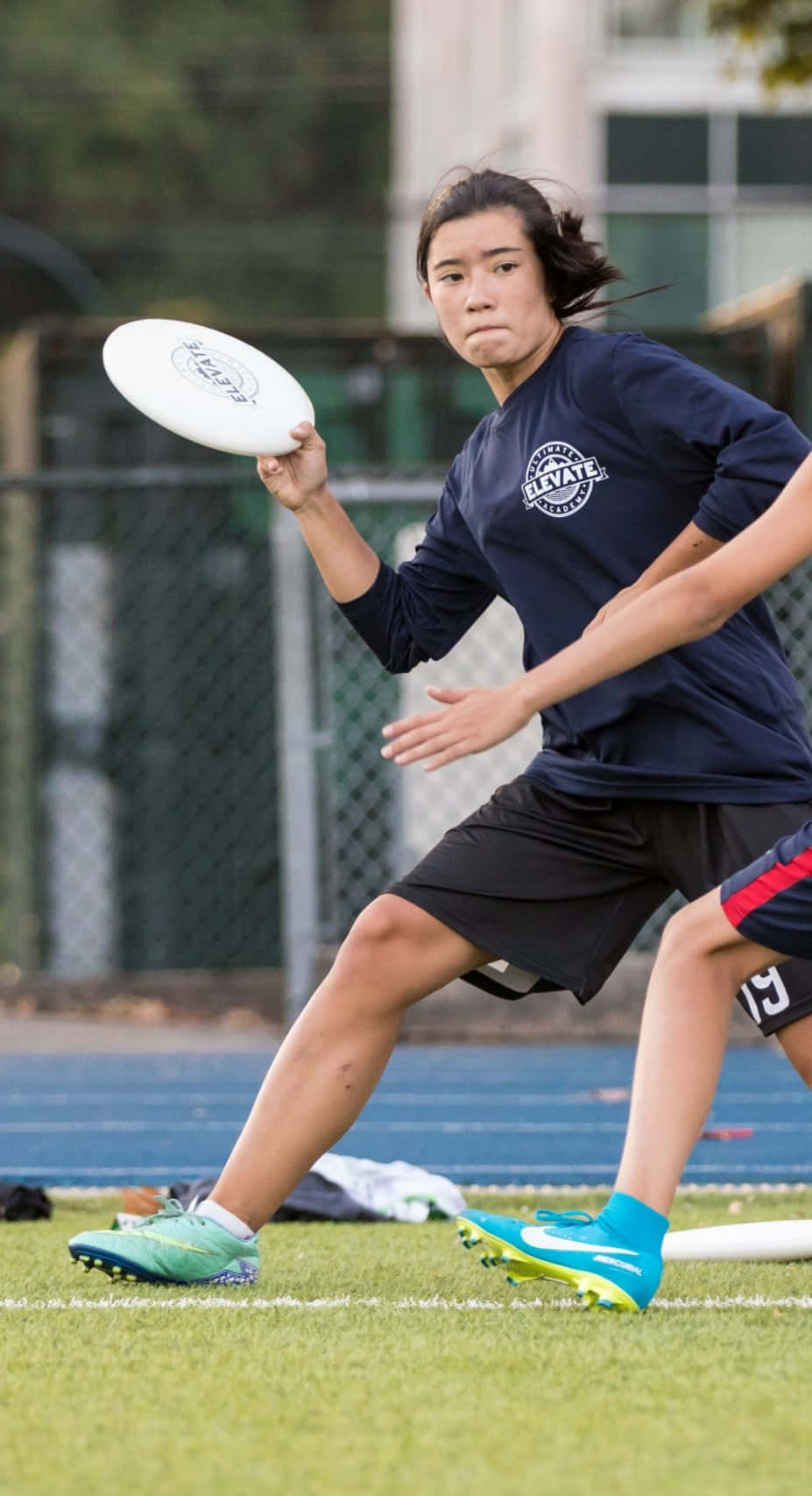 Android Ultimate Frisbee 背景：女運動員投擲飛盤 壁紙 — 一位專注的女運動員穿著海軍藍襯衫和亮藍色鞋子，準備在跑道附近的草地上投擲白色飛盤。