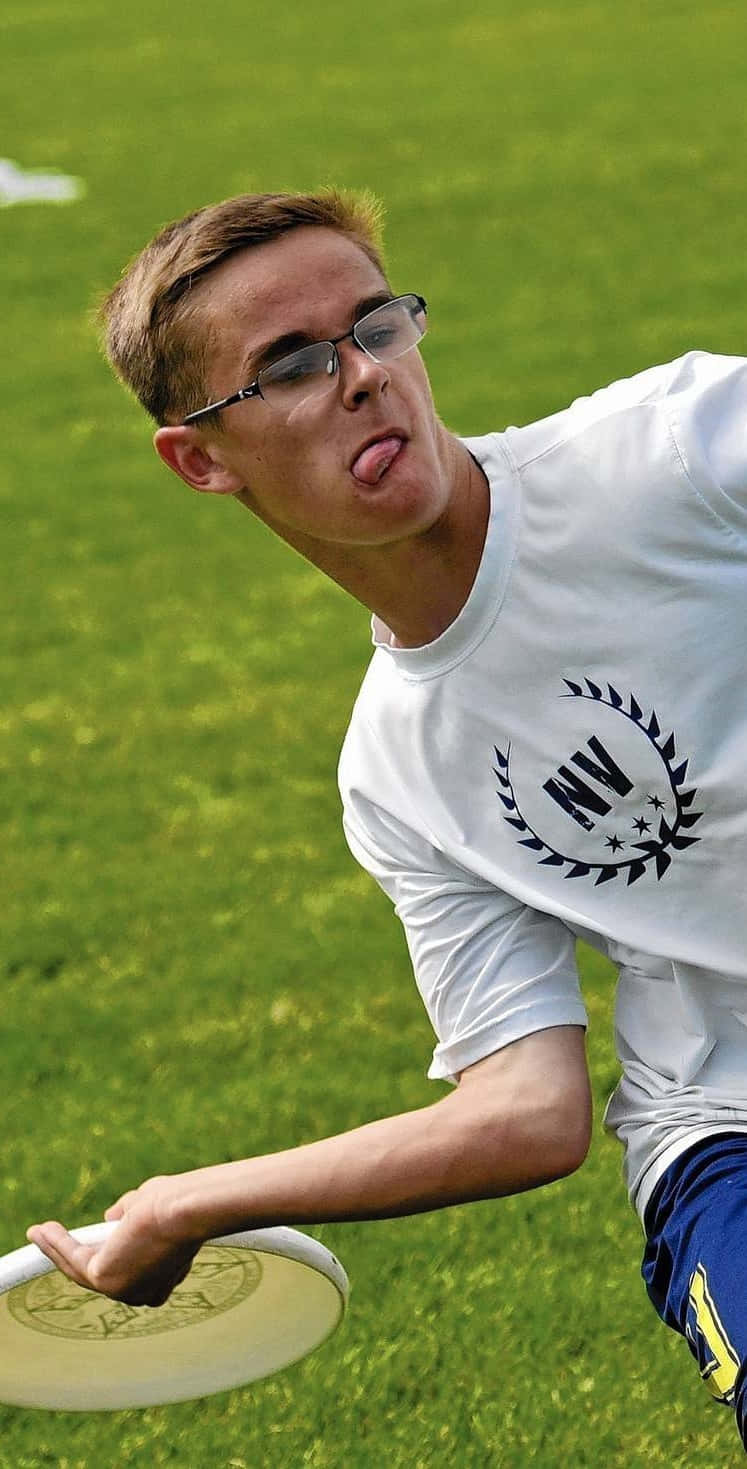 Android Ultimate Frisbee 背景：男運動員搞笑反應 壁紙 — 一個戴眼鏡的年輕男子在充滿活力的綠色場地上抓住飛盤，頑皮地伸出舌頭。