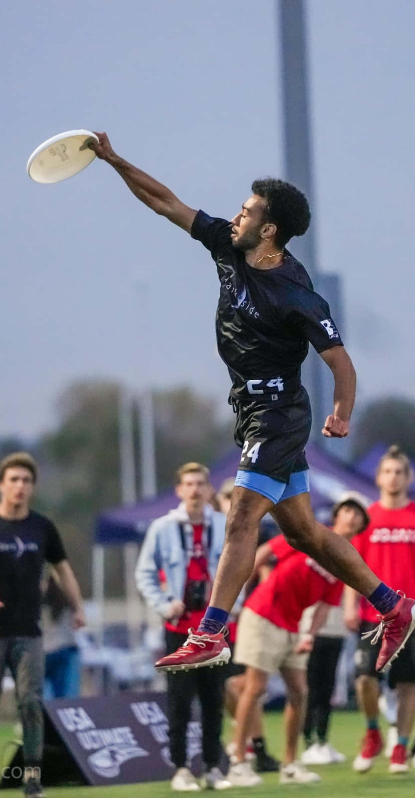 Android Ultimate Frisbee 背景：男運動員接飛盤 壁紙 — 一名男性終極飛盤選手高高躍起，在半空中接住一個白色飛盤，背景是模糊的戶外運動場地。