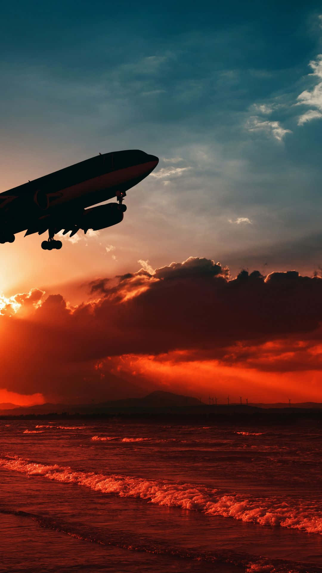 Android 小型飛機翱翔天際 壁紙 — 飛機剪影在火熱的日落海洋上空翱翔，下方有戲劇性的雲彩和發光的波浪。
