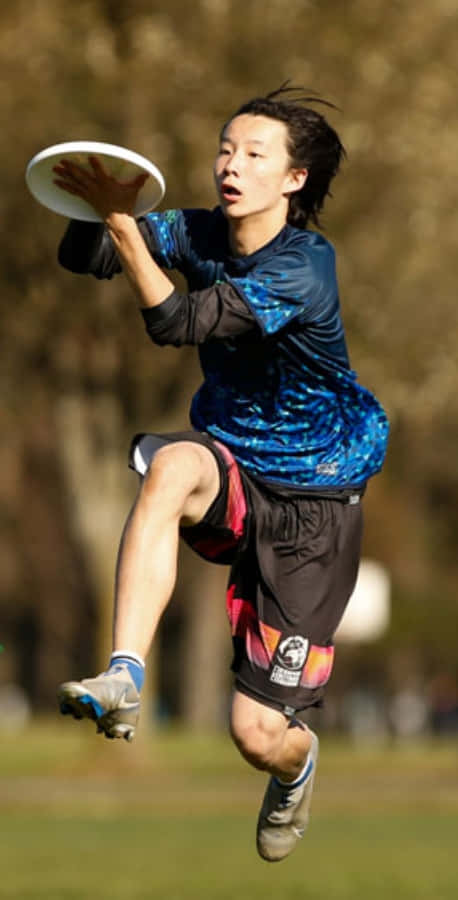 Android手機背景下的Ultimate Frisbee精彩瞬間 壁紙 — 一位年輕運動員在陽光明媚的公園中躍起，在模糊的樹木背景下，專注地接住一個白色飛盤。