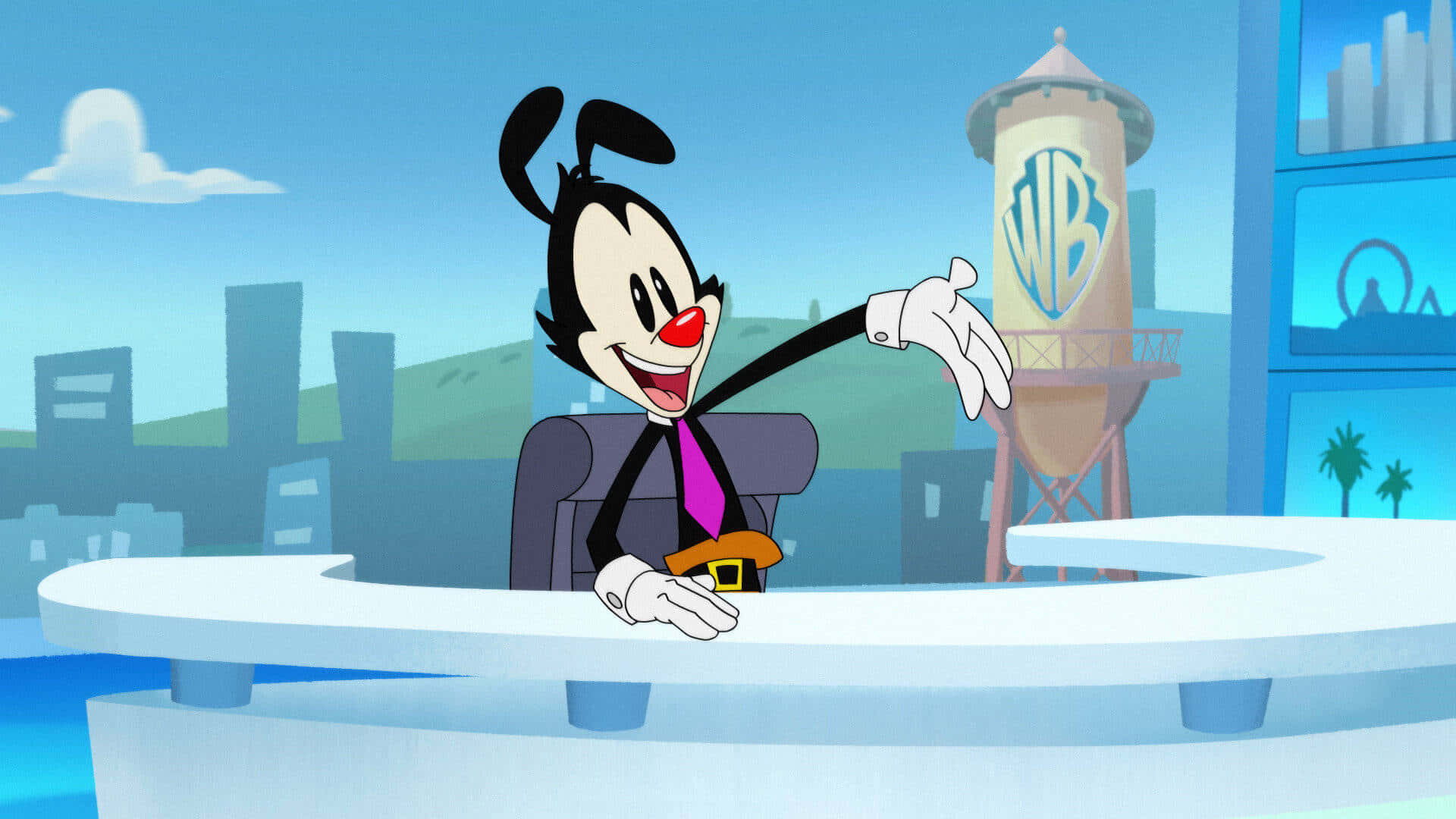 Animaniacs 帶來歡笑與快樂 壁紙 — 一個穿著西裝的開心卡通角色坐在新聞台前，背景有華納兄弟水塔。