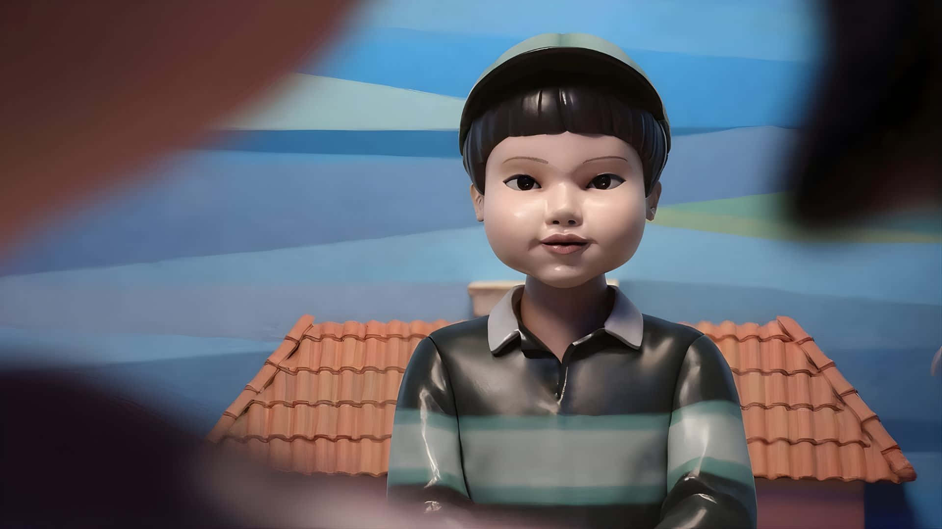  壁紙 — 一個穿著條紋襯衫和帽子的年輕動漫風格男孩站在一個有赤陶屋頂瓦片的風格化房屋前。