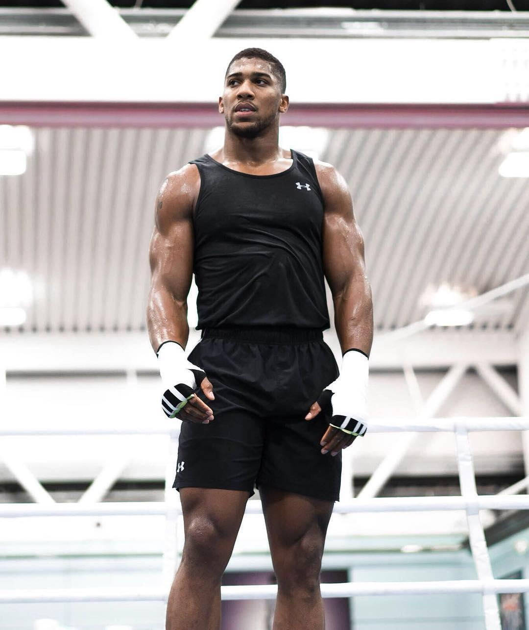 Anthony Joshua 健身房肌肉照 壁紙 — 一位肌肉發達的拳擊手穿著黑色裝備和白色手套，專注地站在燈光明亮的擂台上。