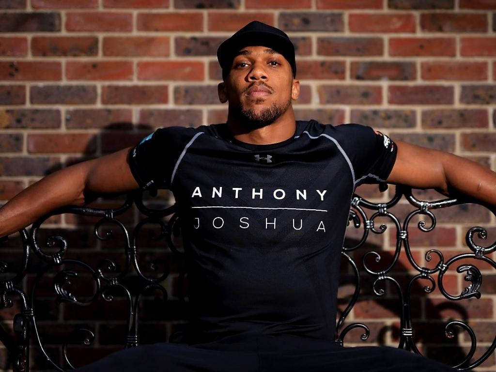 拳擊手 Anthony Joshua 身穿品牌上衣，坐在裝飾長椅上，背景是磚牆。