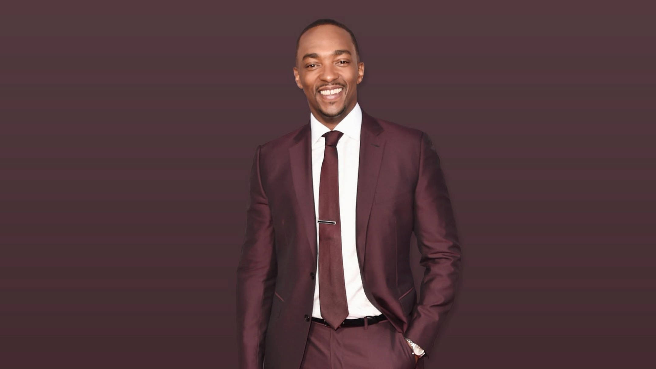 漫威明星 Anthony Mackie 壁紙 — 一個微笑的男人穿著栗色西裝，自信地站在紫色漸變背景前。