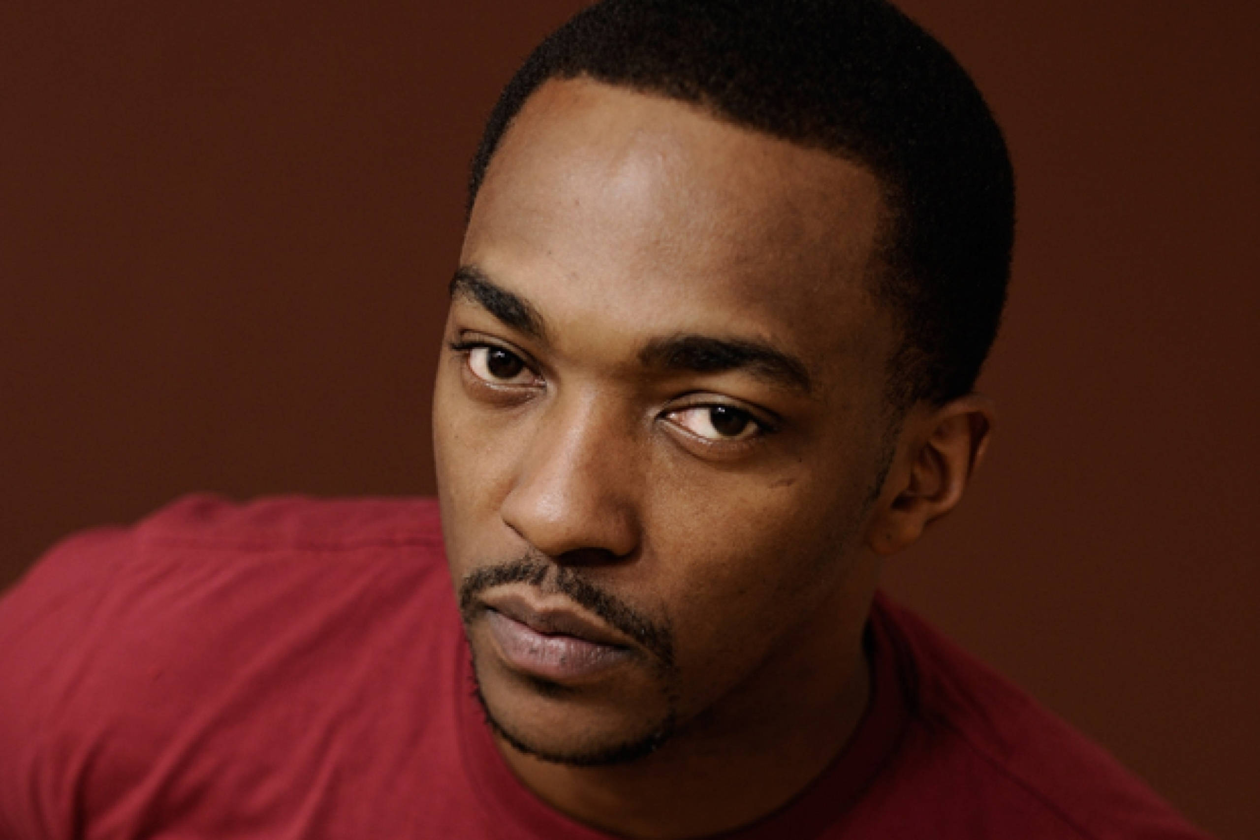 Anthony Mackie 炫目凝視 壁紙 — 一個表情嚴肅嘅男人嘅近距離肖像，佢著住紅色恤衫，背景係溫暖嘅棕色。