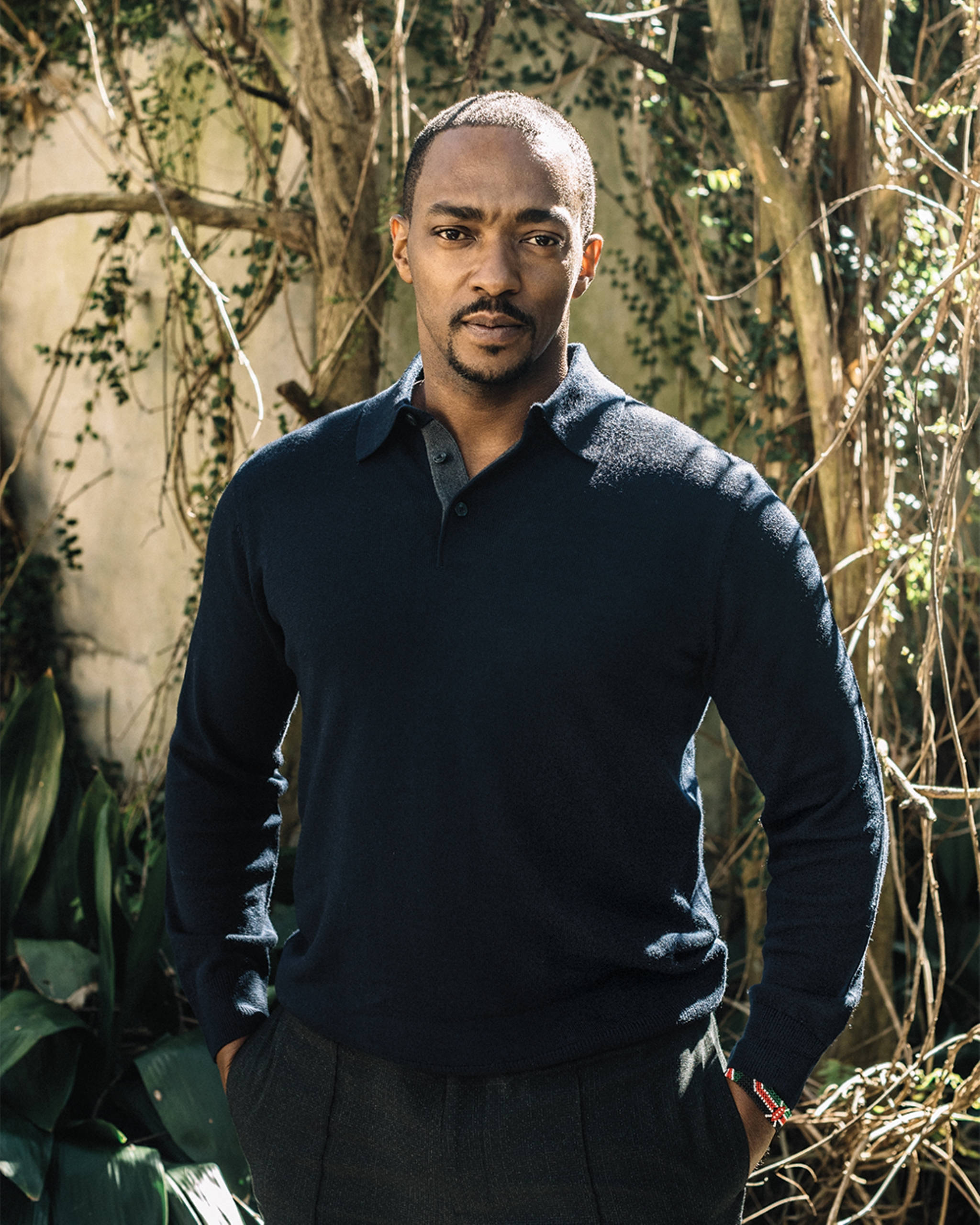 Anthony Mackie 為 Variety 專訪拍攝 壁紙 — 一個穿著深色毛衣的男人自信地雙手插袋，站在陽光普照的花園背景下，身後有藤蔓和樹木。