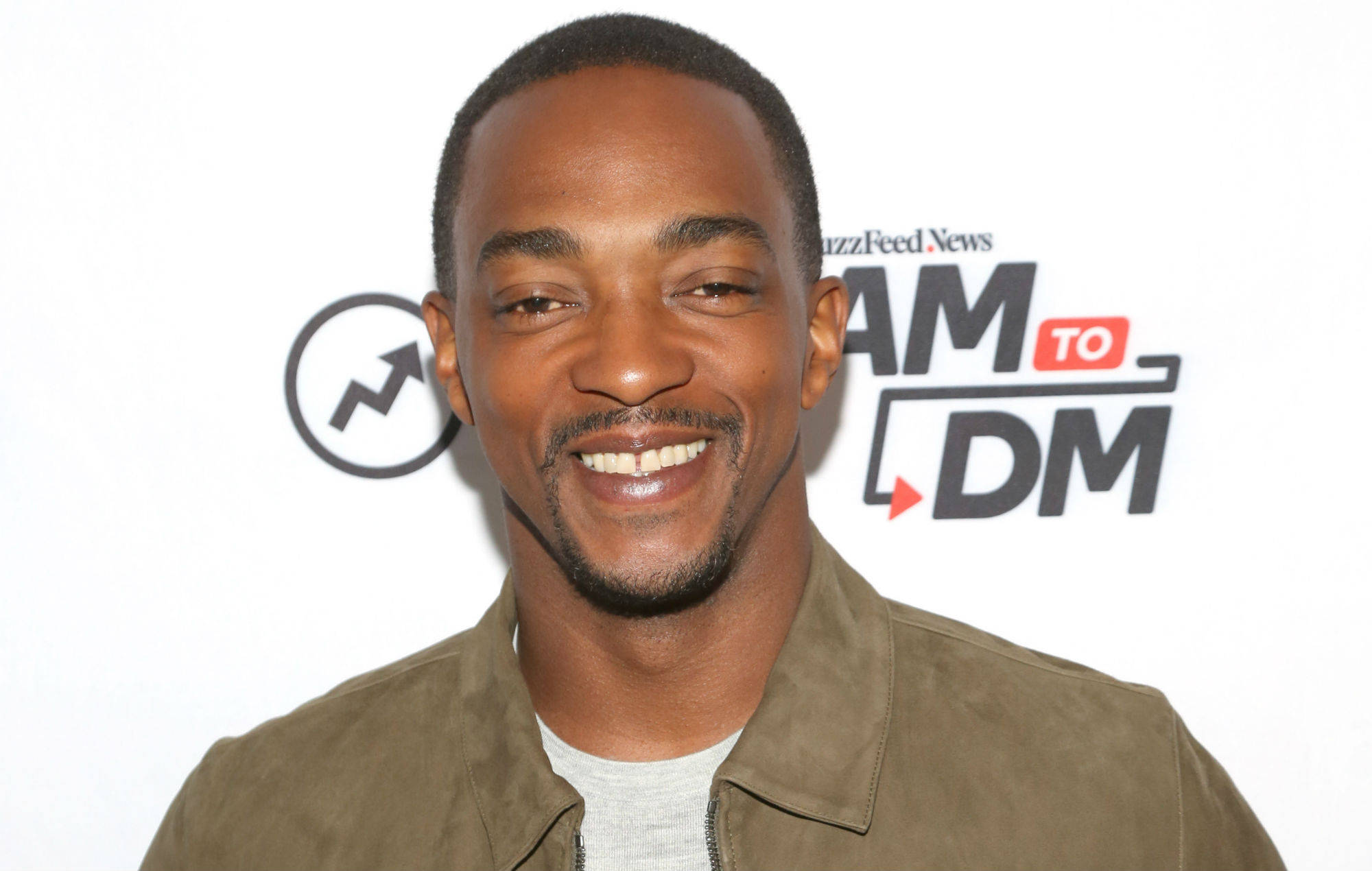 Anthony Mackie 獨特微笑 壁紙 — 一名面帶微笑、身穿棕色麂皮外套的男子站在白色背景前，背景上有Buzzfeed News和AM to DM標誌。