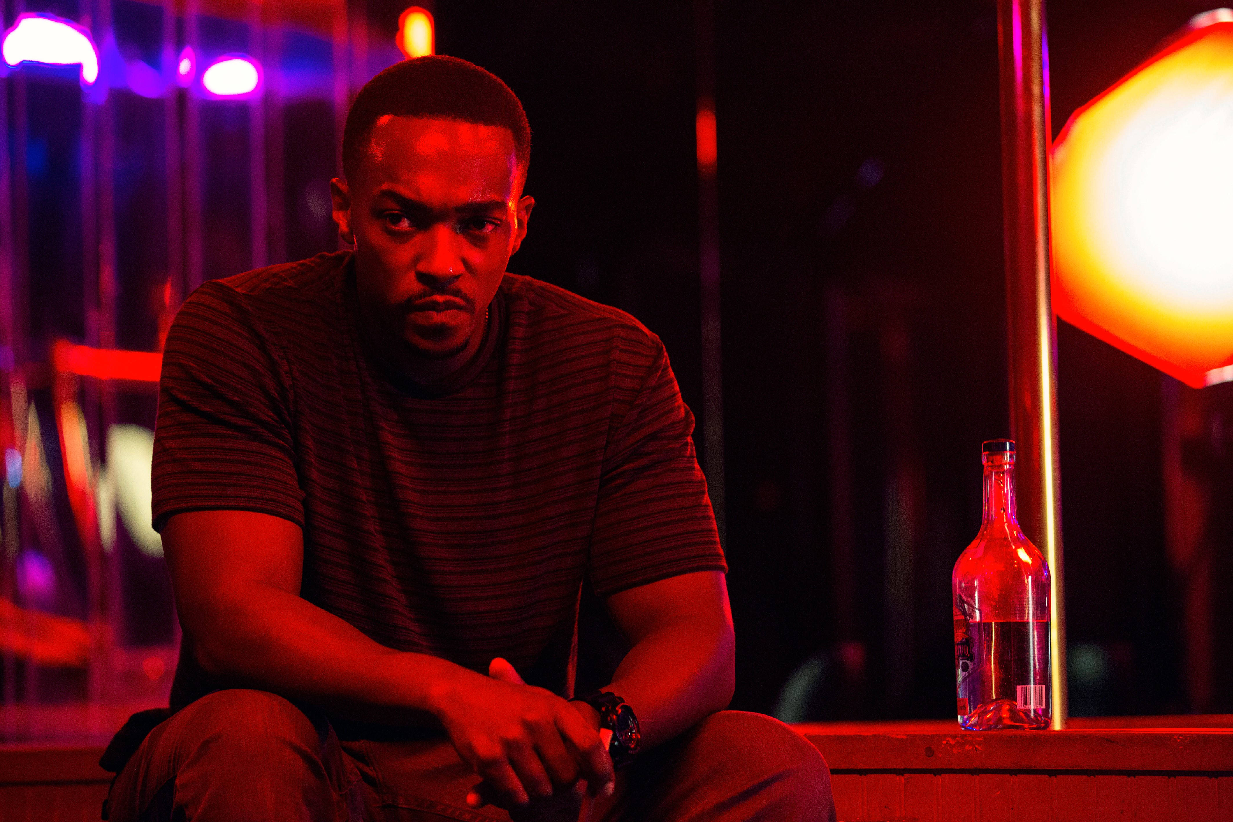 Anthony Mackie 飾 Marcus Belmont 壁紙 — 一個男人獨自坐在昏暗的酒吧裡，被紅色和紫色的霓虹燈光包圍，旁邊放著一個瓶子。