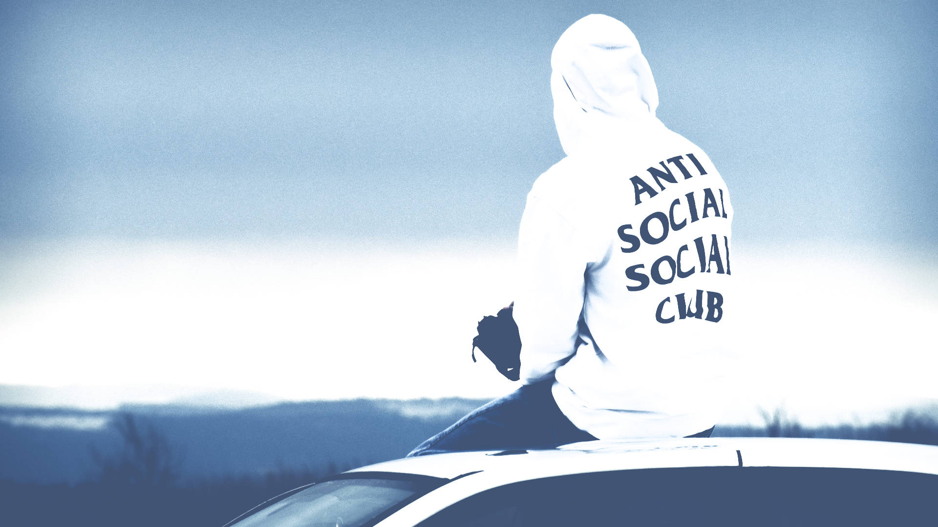 Anti Social Social Club 女裝連帽衫 壁紙 — 一個人穿著白色連帽衫，上面有「ANTI SOCIAL SOCIAL CLUB」字樣，坐在一輛汽車上，背景是朦朧的藍天。