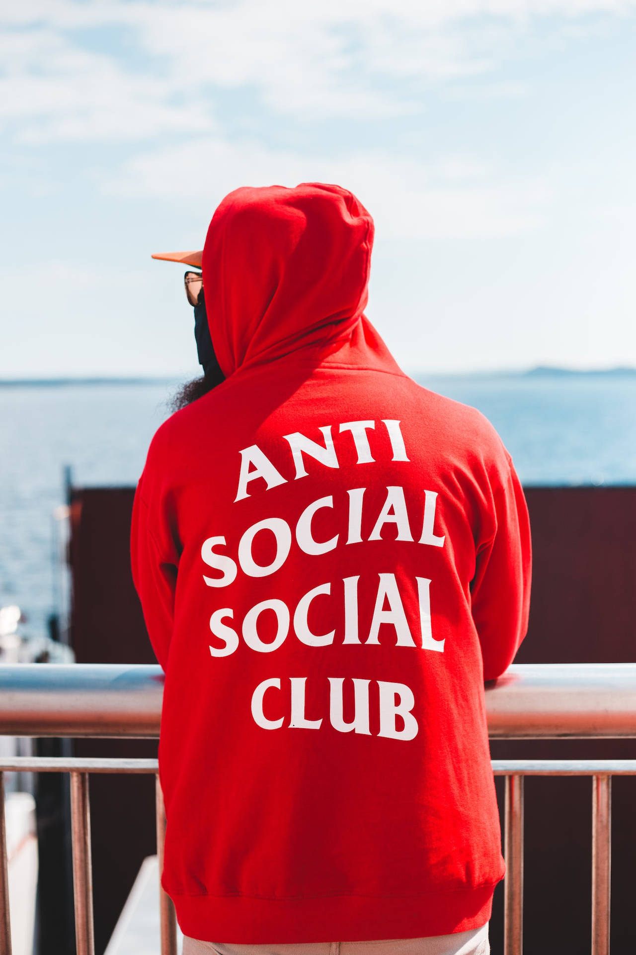 Anti Social Social Club紅色連帽衫 壁紙 — 一個穿著印有「ANTI SOCIAL SOCIAL CLUB」字樣紅色連帽衫的人，站在欄杆上俯瞰晴朗天空下的海洋。