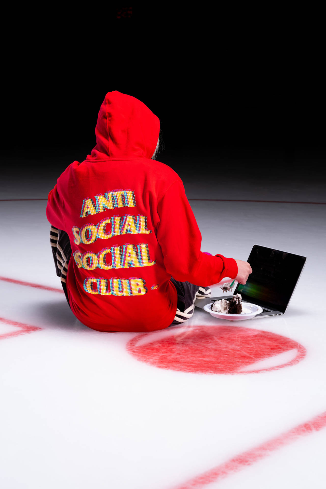 Anti Social Social Club連帽衫與蛋糕 壁紙 — 一個穿著紅色連帽衫的人坐在有紅色標記的白色地板上，一邊吃蛋糕一邊使用筆記型電腦，背景為黑色。