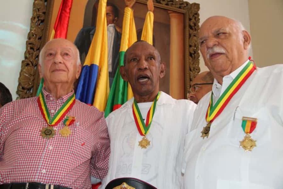 三位拳壇傳奇：Antonio Nader、Antonio Cervantes 與 Alfonso Múnera 壁紙 — 三位身穿白恤衫的長者佩戴著紅、黃、綠絲帶的獎章，站在旗幟和裱框肖像前。