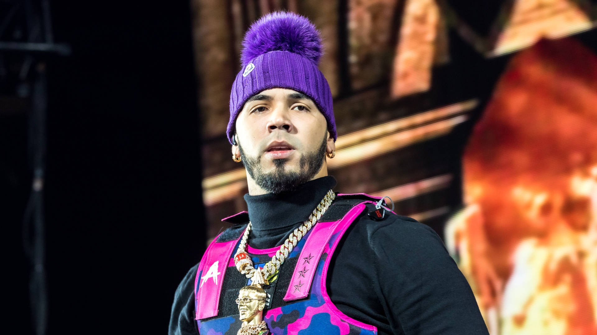 Anuel AA《Intocable》演出 壁紙 — 一個戴著紫色絨球冷帽和金鏈的男人，在火熱的舞台背景下凝視著。