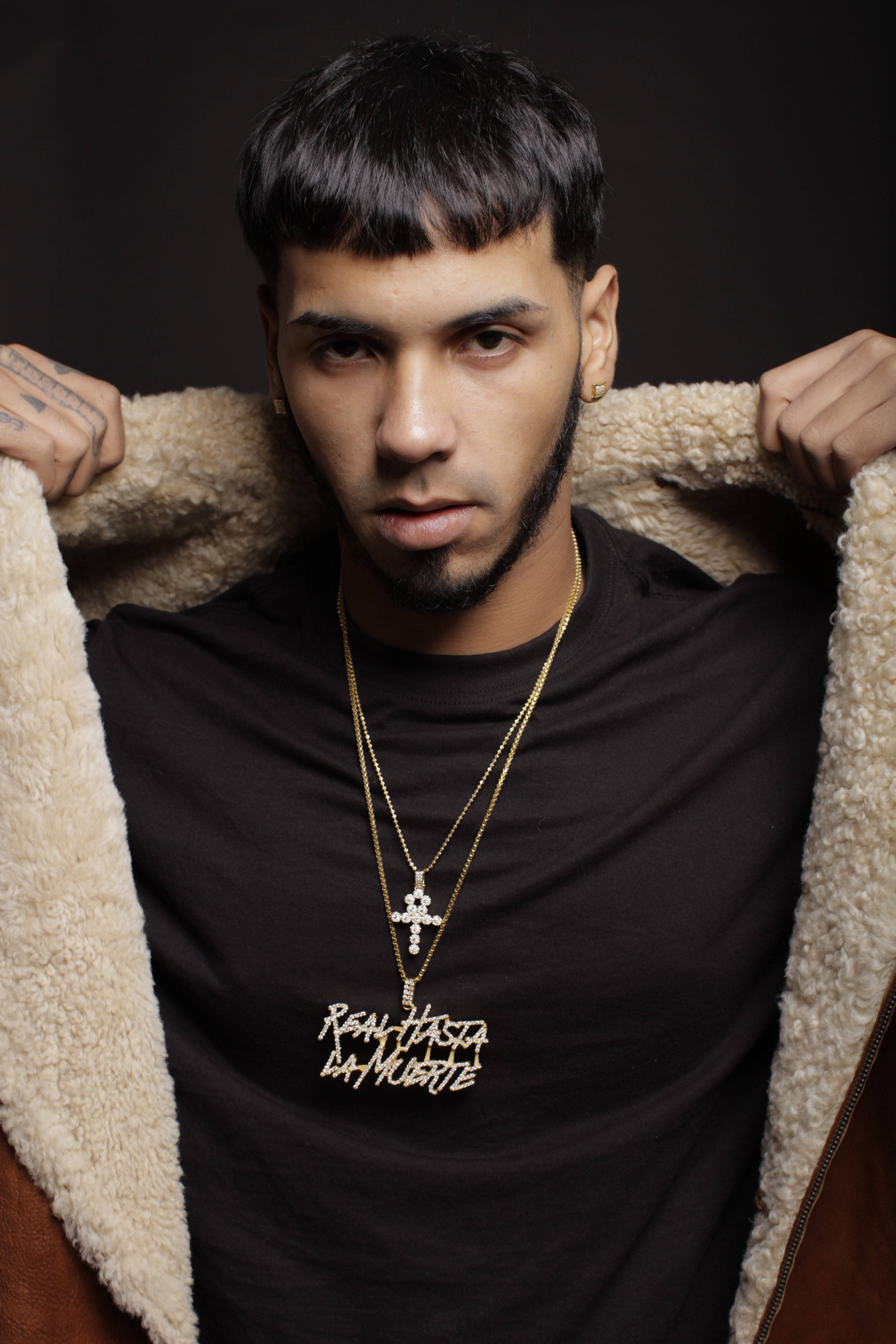 Anuel AA《Real Hasta La Muerte》造型 壁紙 — 一個留鬍鬚和有紋身的男人拉開一件羊羔毛外套，露出黑色T恤，上面有金鏈和十字架吊墜。