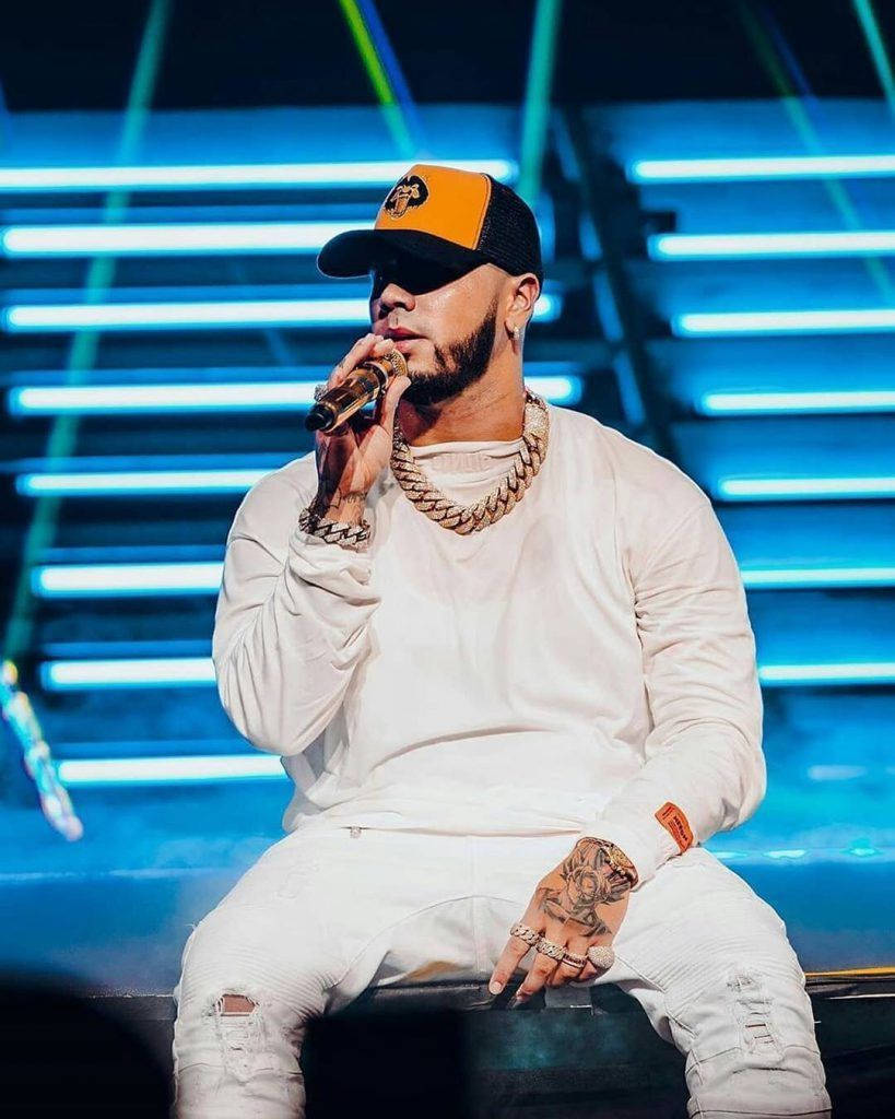 Anuel AA 全白造型 壁紙 — 一位身穿白色服裝、戴橙色帽子的說唱歌手在藍色霓虹燈照亮的舞台上表演。