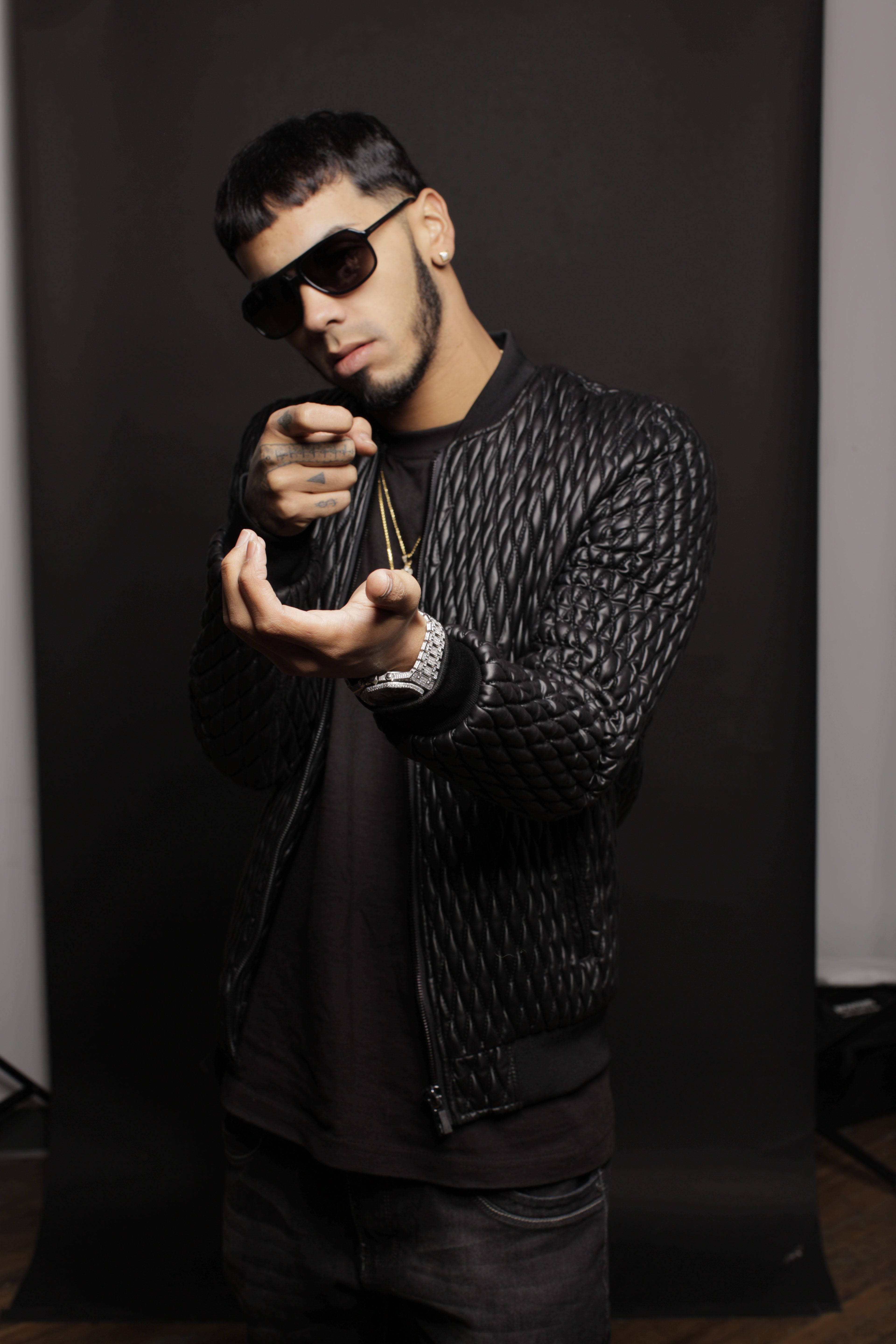 Anuel AA 全黑造型 壁紙 — 一位時尚男士穿著黑色絎縫外套和太陽眼鏡，在深色工作室背景前自信地擺姿勢。