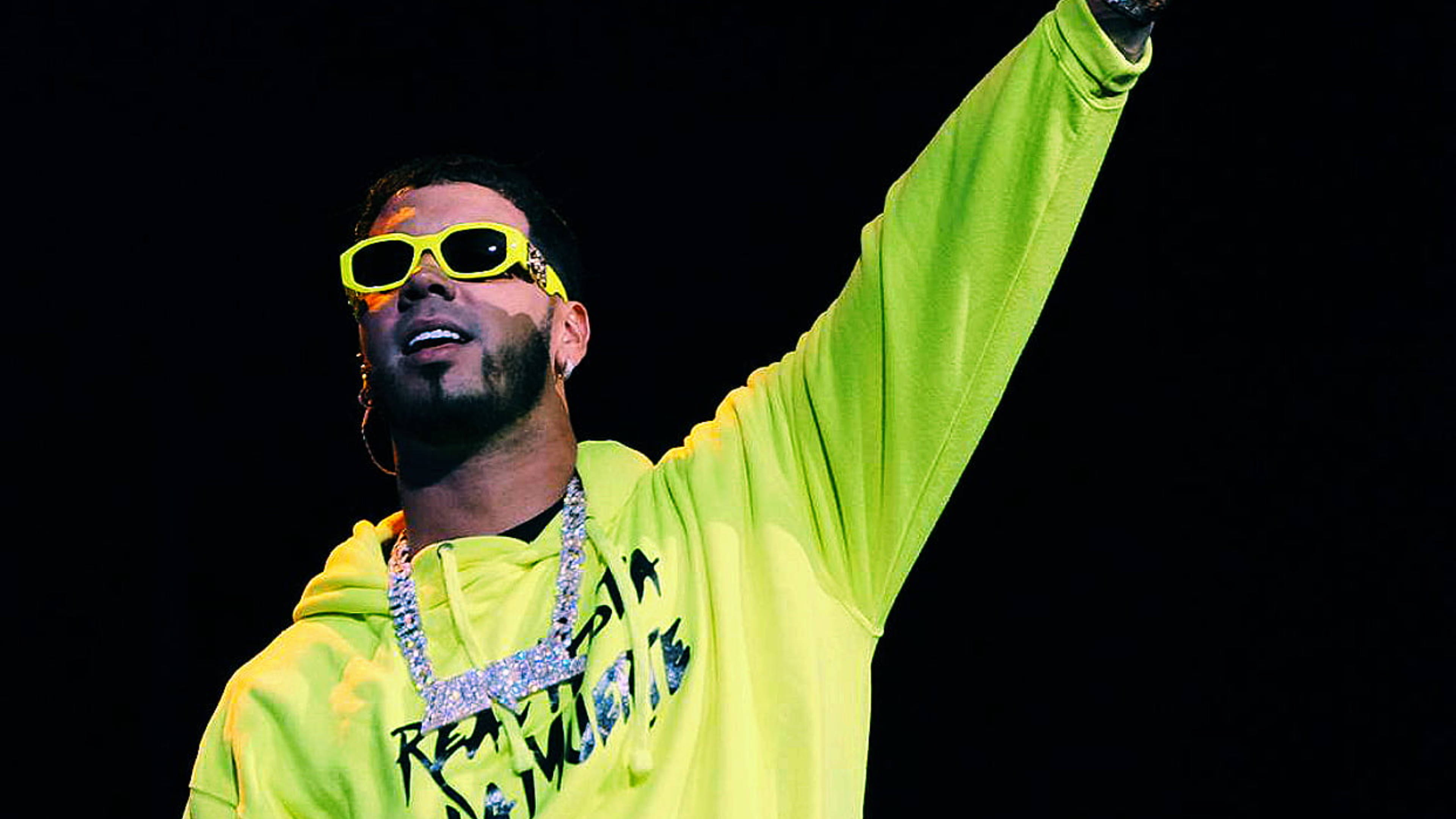 Anuel AA 巡迴演唱會舞台表演 壁紙 — 一位表演者身穿螢光黃色連帽衫和相配的太陽眼鏡，在黑色背景下勝利地舉起一隻手臂。