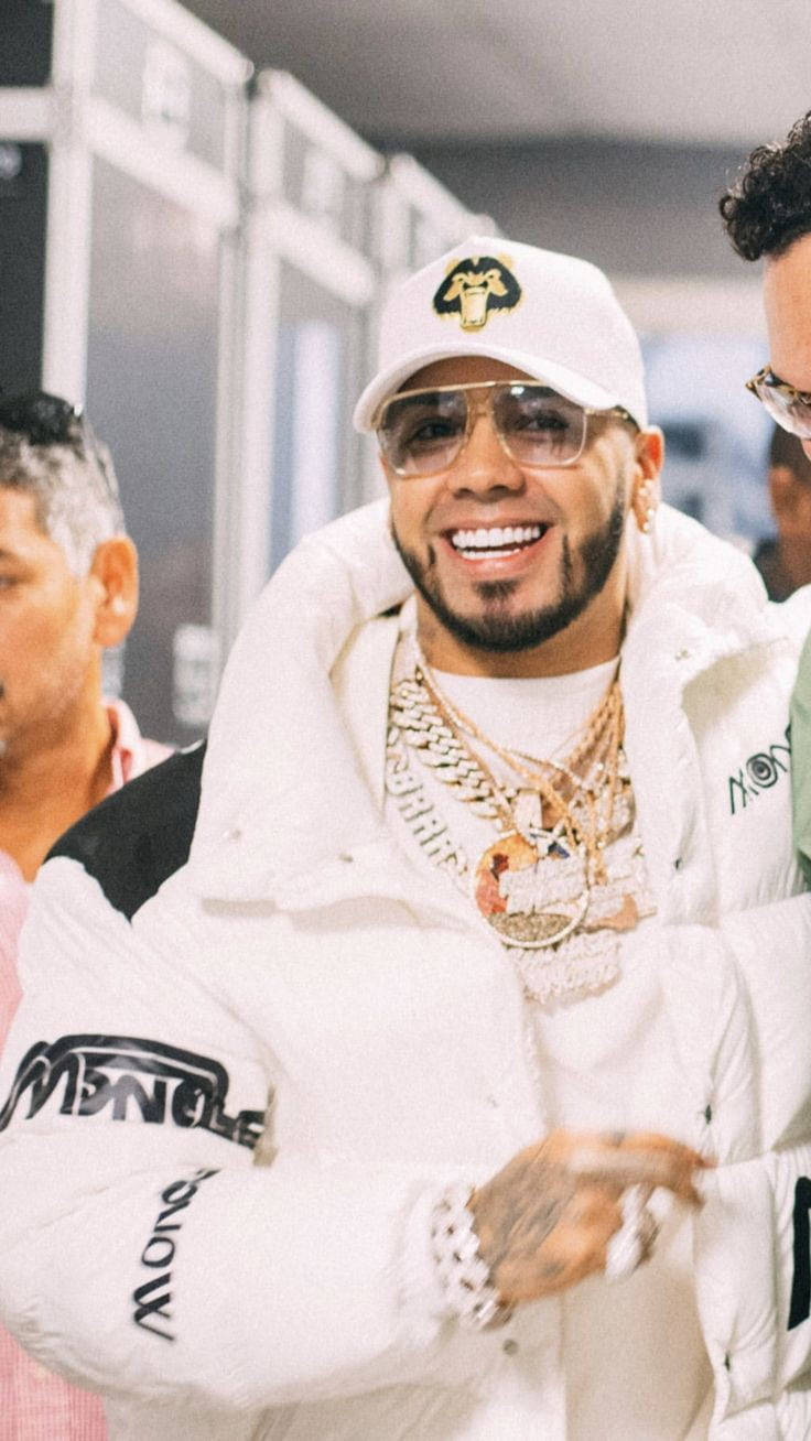 Anuel AA 拉丁歌手與饒舌歌手 壁紙 — 一位微笑的男子穿著白色Moncler外套和金色鏈條，自信地在室內與其他人一起擺姿勢。