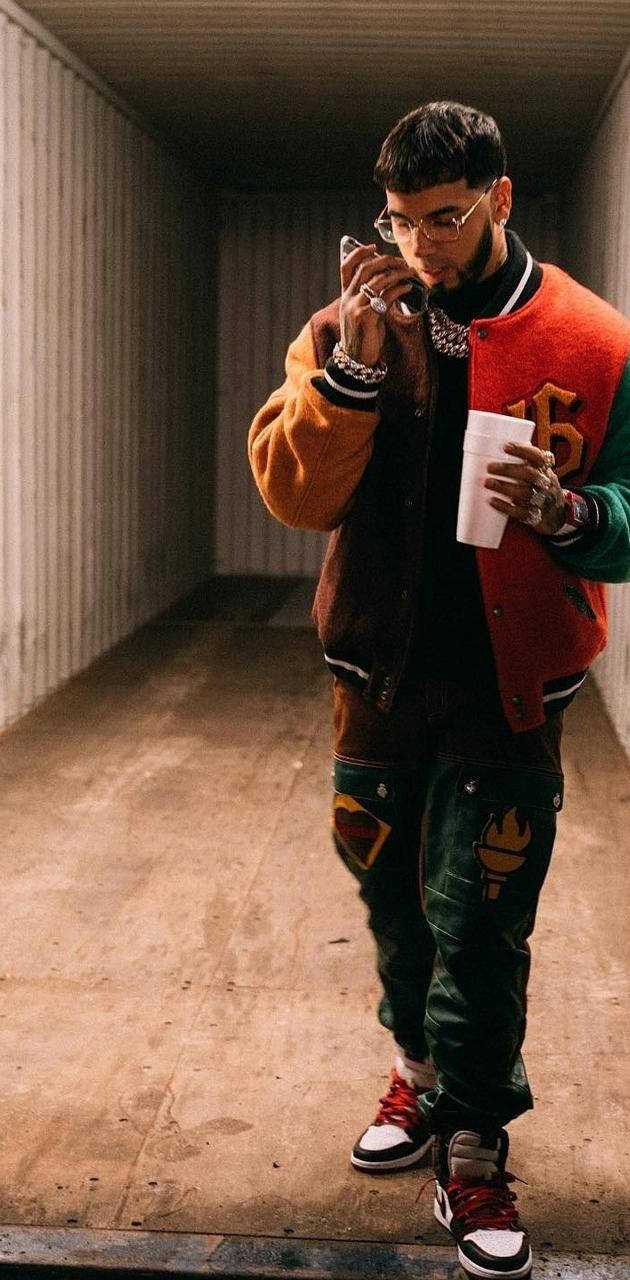 Anuel AA 時尚造型 壁紙 — 一位時尚的年輕男子穿著色彩繽紛的棒球外套和圖案褲，手持電話和咖啡杯，穿過狹窄的工業走廊。