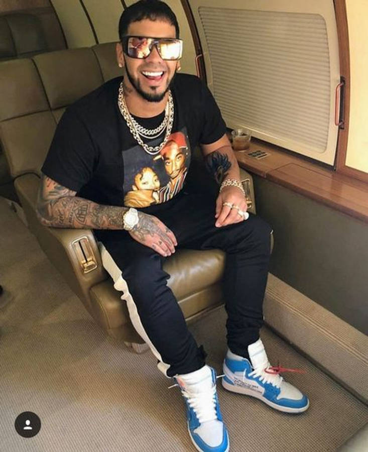 Anuel AA 時尚隨拍 壁紙 — 一個微笑的男人，身上有紋身，戴著反光太陽眼鏡，自信地坐在私人飛機上，穿著藍白運動鞋。