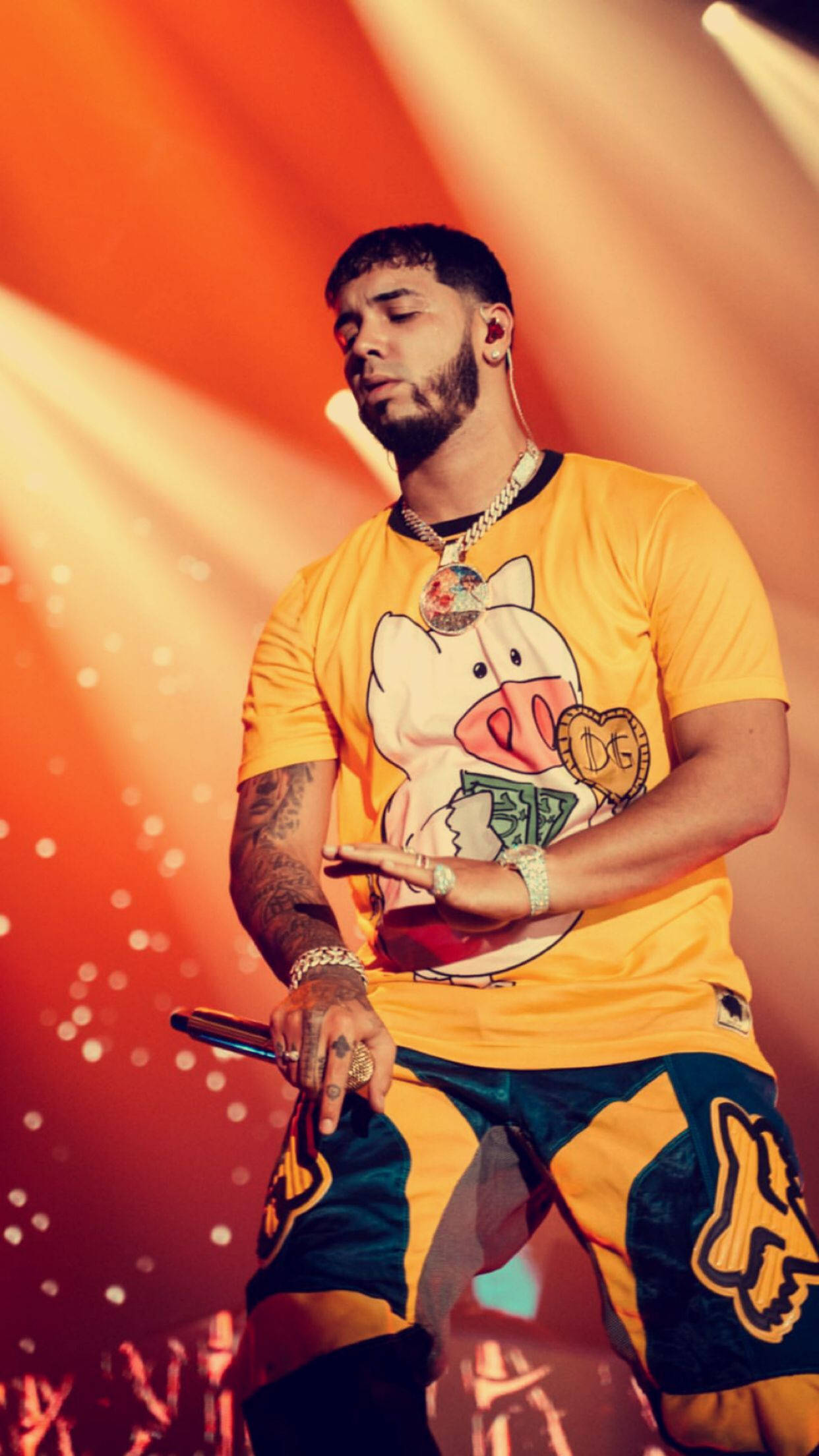 Anuel AA 橙色造型 壁紙 — 一位身穿亮黃色襯衫（帶有豬圖案）的男性表演者站在舞台上，在溫暖的橙色聚光燈下拿著麥克風。