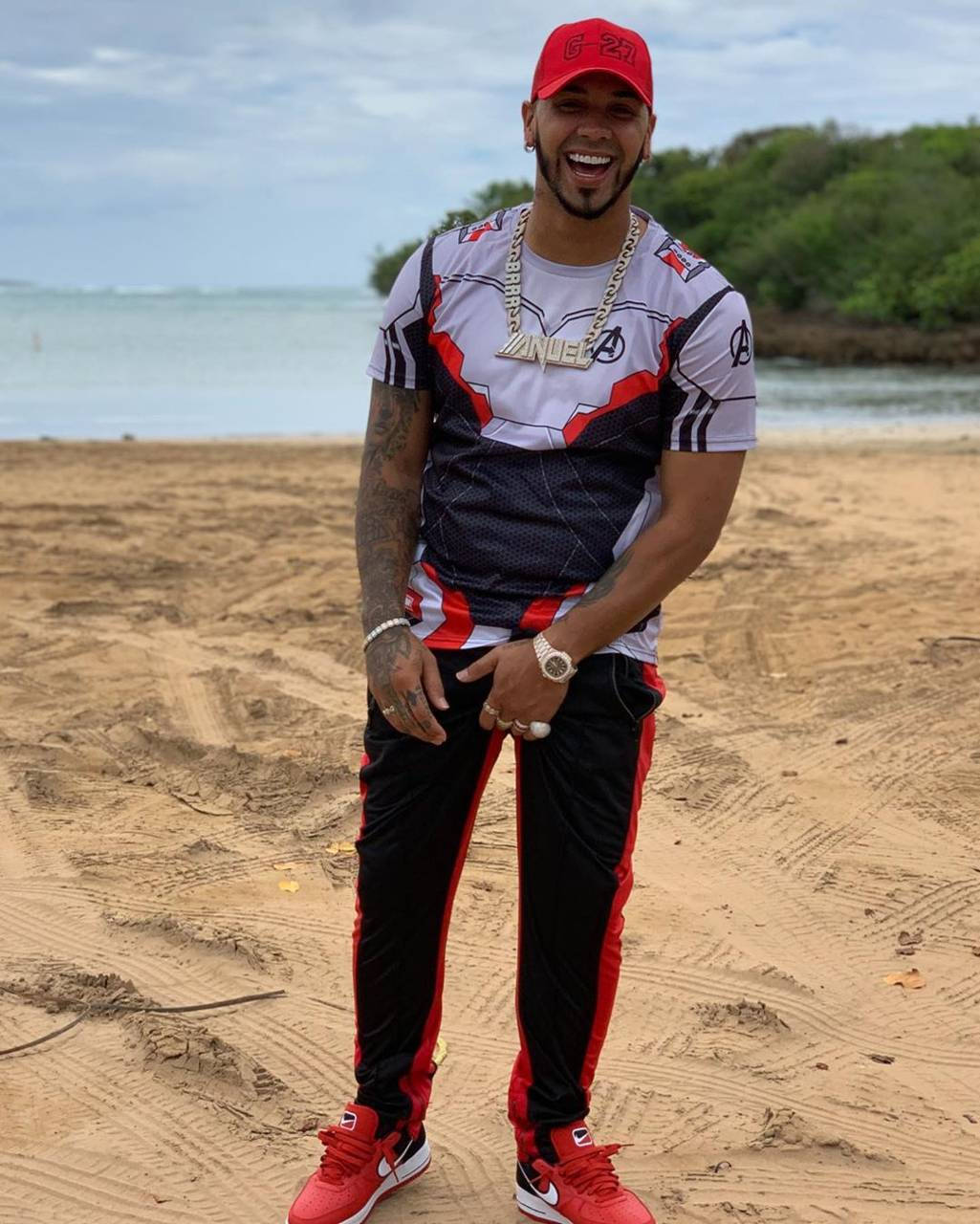 Anuel AA 海灘時刻 壁紙 — 一個戴紅帽、穿復仇者聯盟T恤的微笑男子站在沙灘上，背後是海洋和綠植。