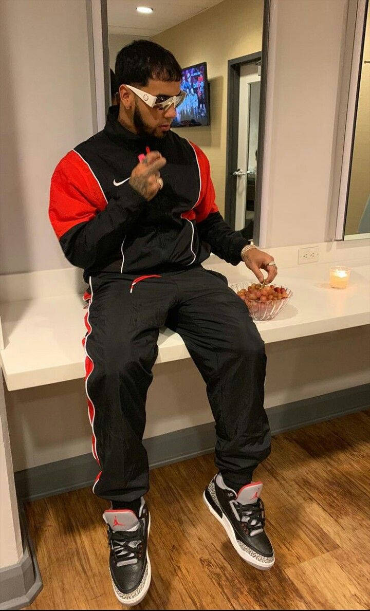 Anuel AA 真實瞬間 壁紙 — 一名身穿黑色和紅色運動服的男子隨意地坐在白色櫃檯上，戴著白色太陽眼鏡吃零食。