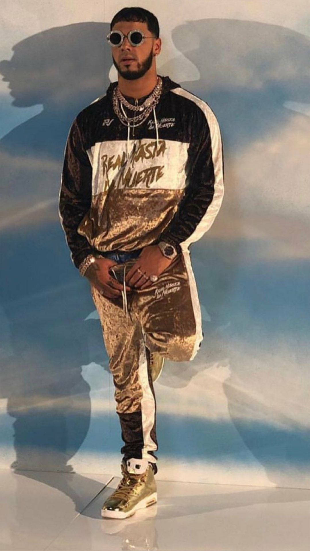 Anuel AA 美學風格 壁紙 — 一名身穿金色和黑色天鵝絨運動服、戴著太陽眼鏡的男子自信地站在多雲的天空背景下。