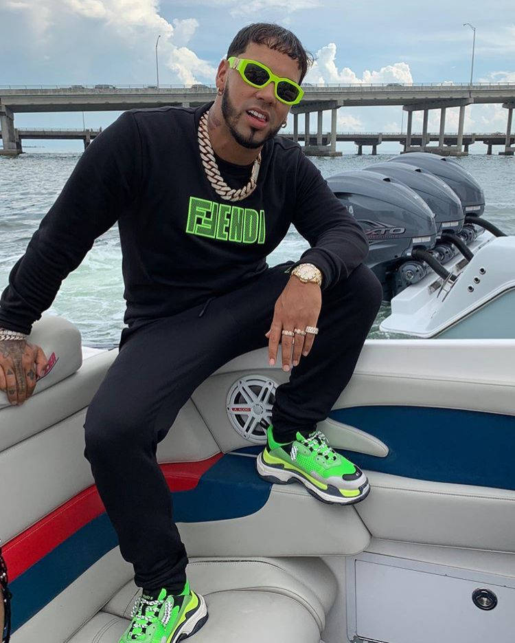 Anuel AA 與 Fendi 壁紙 — 一位時尚男士戴著螢光綠色太陽眼鏡和運動鞋，在快艇上自信地擺姿勢，背景是一座橋樑。