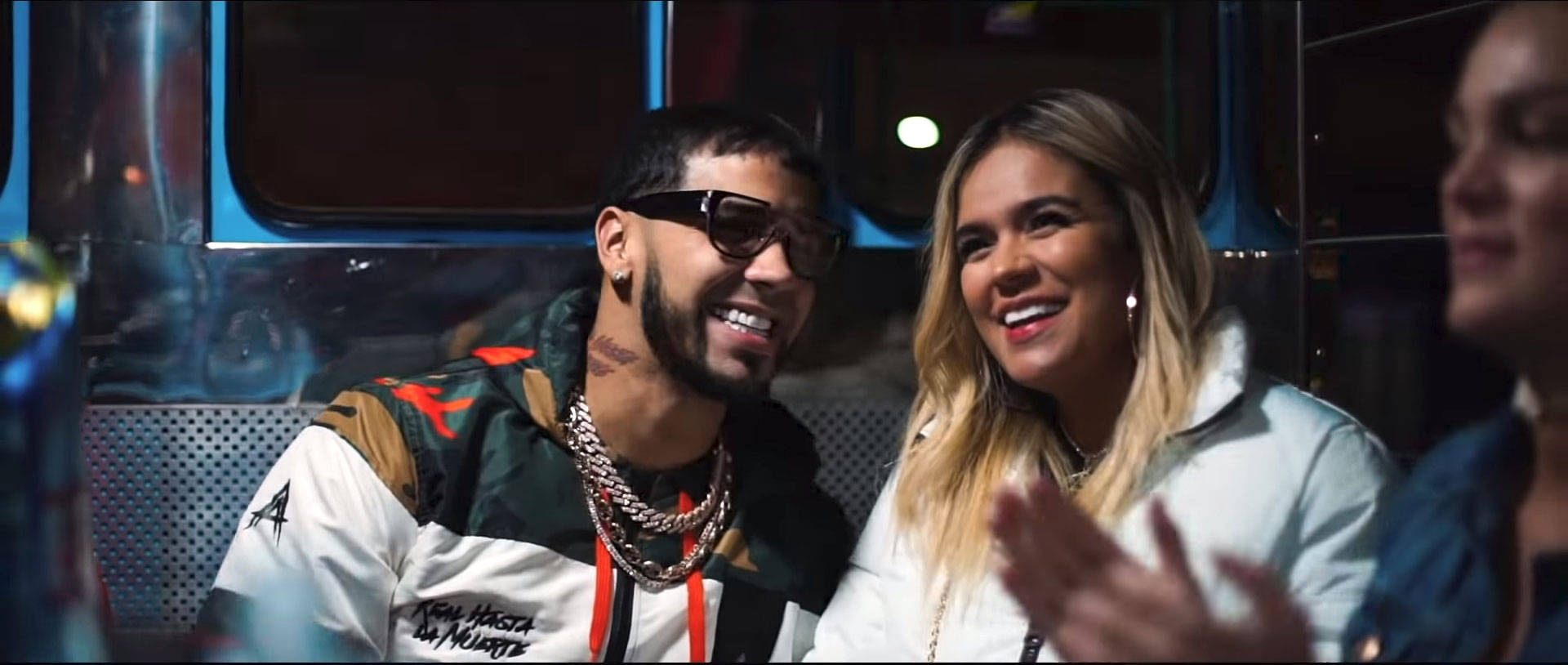 Anuel AA 與 Karol G 壁紙 — 兩個微笑的人在昏暗的車輛中，穿著鮮豔的衣服，在溫暖的燈光下分享快樂的時刻。