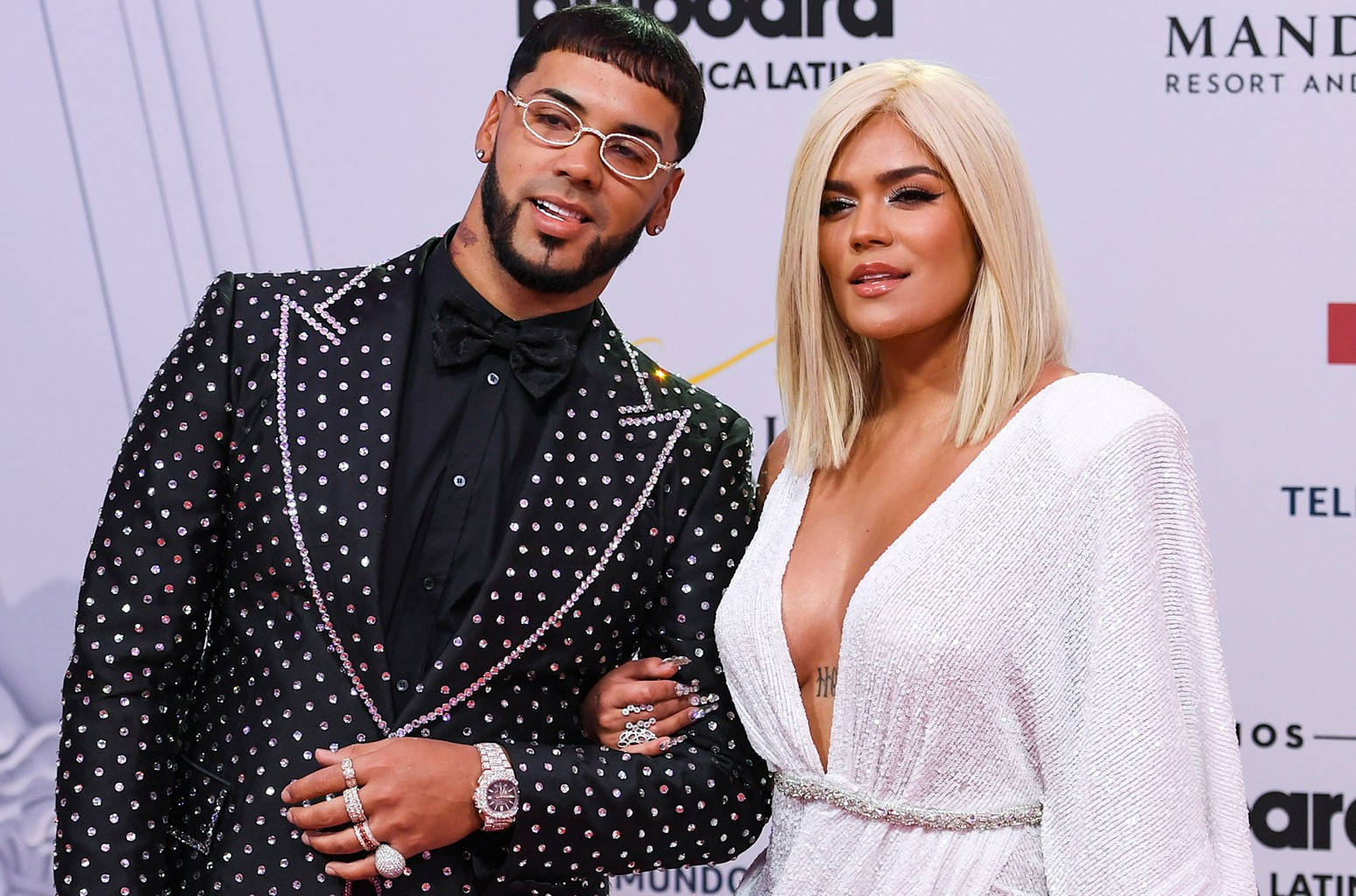 Anuel AA 與 Karol G 榜單頒獎禮 壁紙 — 一名身穿黑色圓點西裝的男子和一名身穿白色禮服的女子在有品牌背景的紅地毯活動中一起擺姿勢。