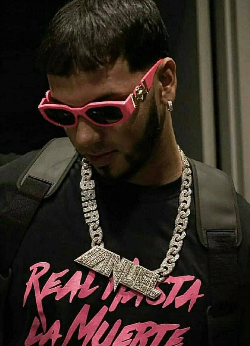 Anuel AA 近照 壁紙 — 一個留鬍鬚的男人戴著大膽的粉紅色太陽眼鏡，並在黑色T恤上戴著一條鑲有鑽石的鏈條，T恤上有粉紅色文字。