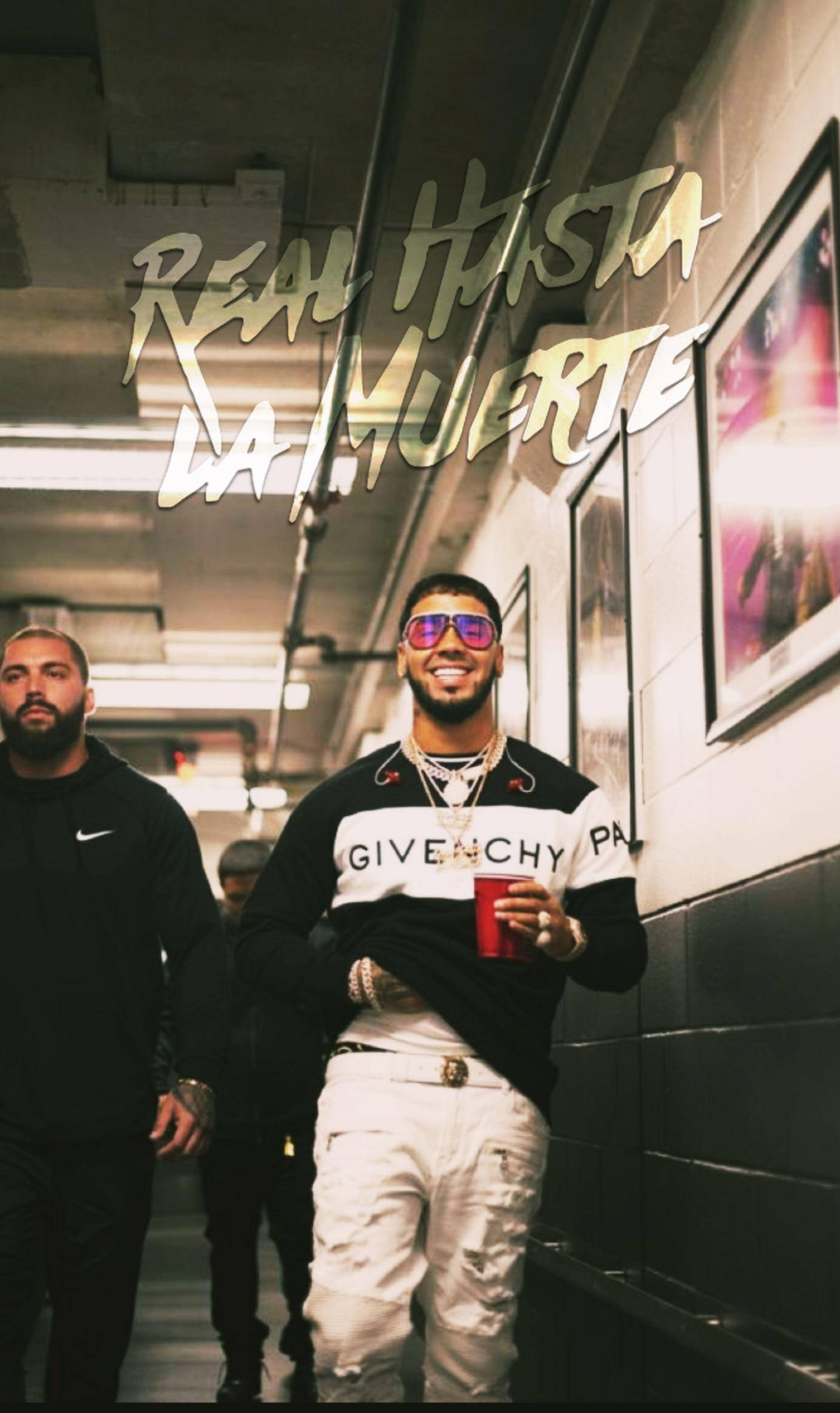 Anuel AA 雷鬼頓粉絲藝術海報 壁紙 — 一位時尚男士穿著白色褲子和Givenchy襯衫，微笑著在昏暗的走廊裡拿著紅色杯子，頭頂有塗鴉文字。