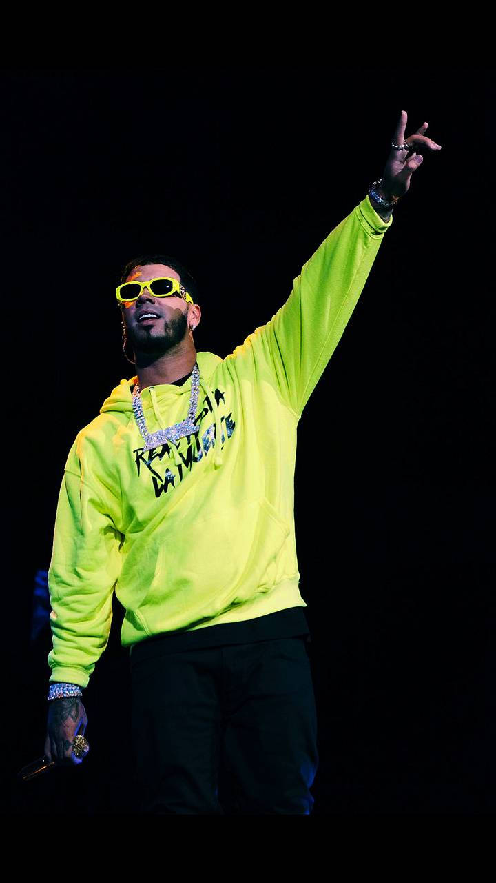 Anuel AA 霓虹綠夾克 壁紙 — 一位表演者穿著螢光黃色連帽衫和太陽眼鏡，在黑色背景上勝利地舉起一隻手臂。