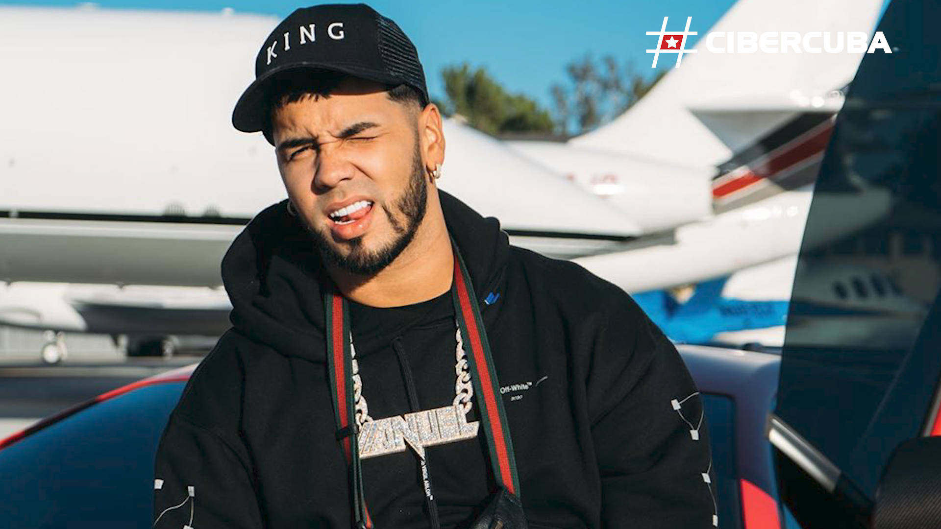 Anuel AA 饒舌歌手 壁紙 — 一個戴著黑色「KING」帽和連帽衫的男人在陽光明媚的停機坪上站在私人飛機旁邊，做鬼臉。